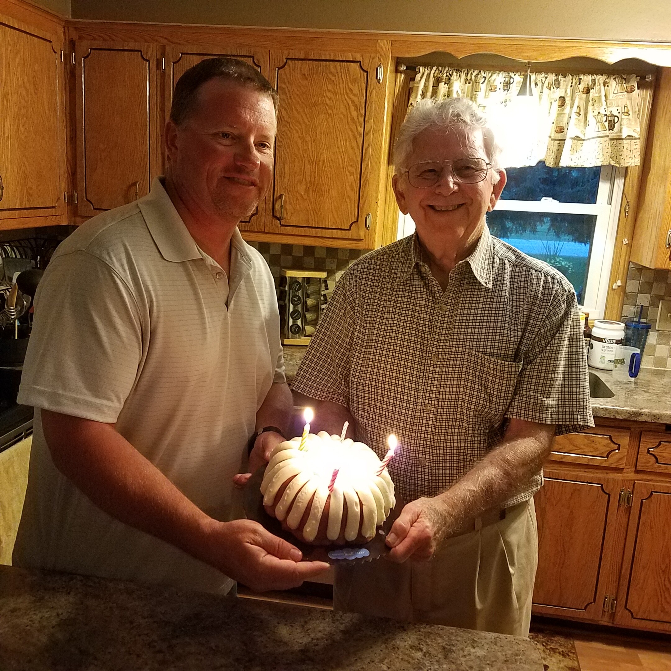 Two men in a kitchen smile as they cut or present a white frosted birthday cake with lit candles.