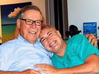 Older man in a blue shirt and glasses hugs a smiling younger man in teal scrubs, both happy in a casual setting.