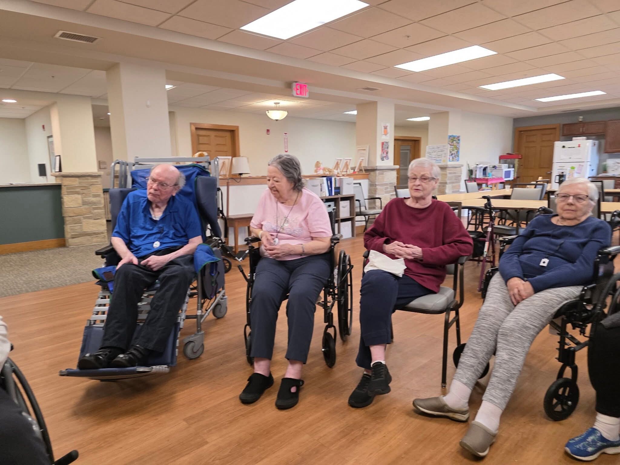 Group of elderly adults in wheelchairs seated in a bright, open common room with wood floors and ceiling lights.