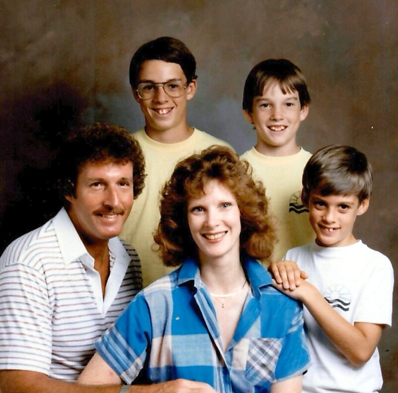 Family portrait of five smiling people posed together in a studio backdrop, likely a parent couple with three children behind them.