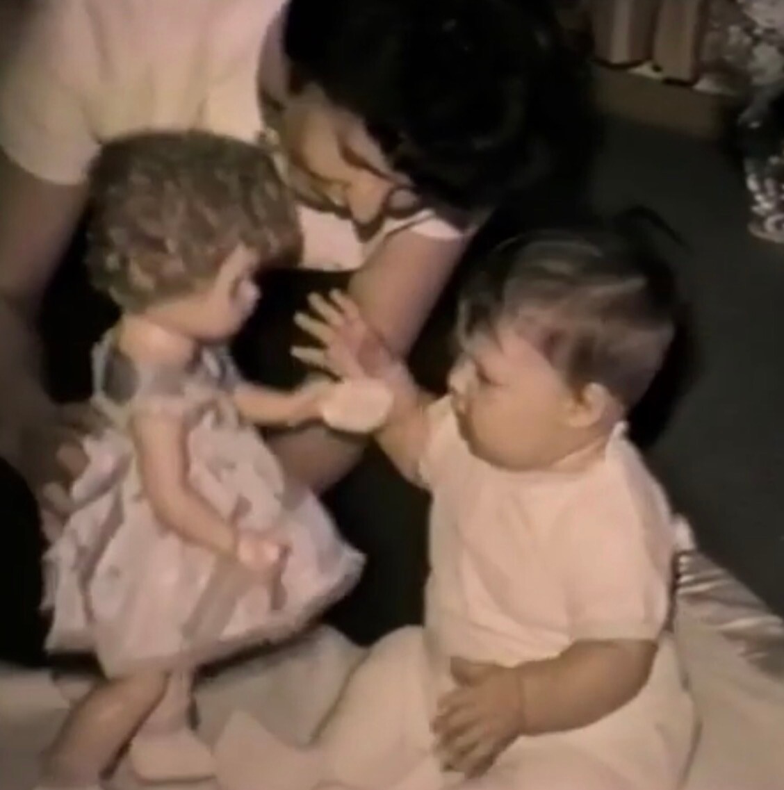 Two toddlers in light outfits reach toward each other and touch hands as an adult looks on.