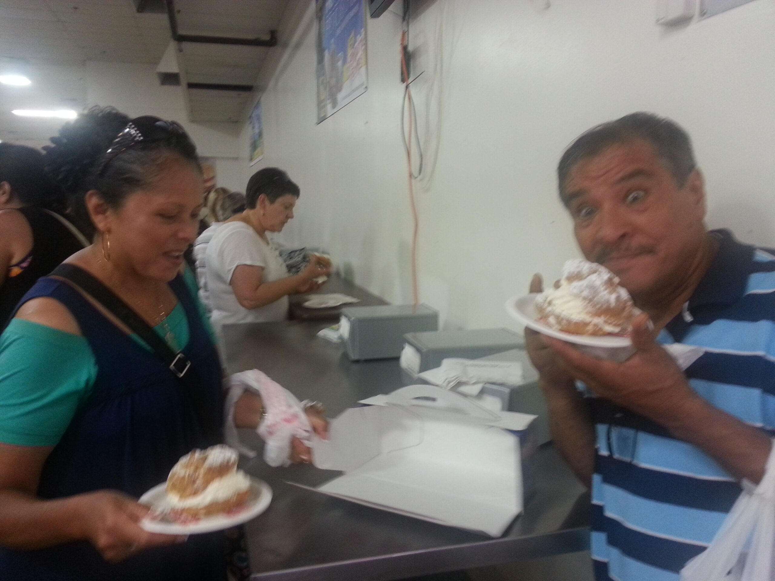 People in a casual shop line holding plates with ice cream cones; a man in a blue striped shirt smiles at the camera on the right.