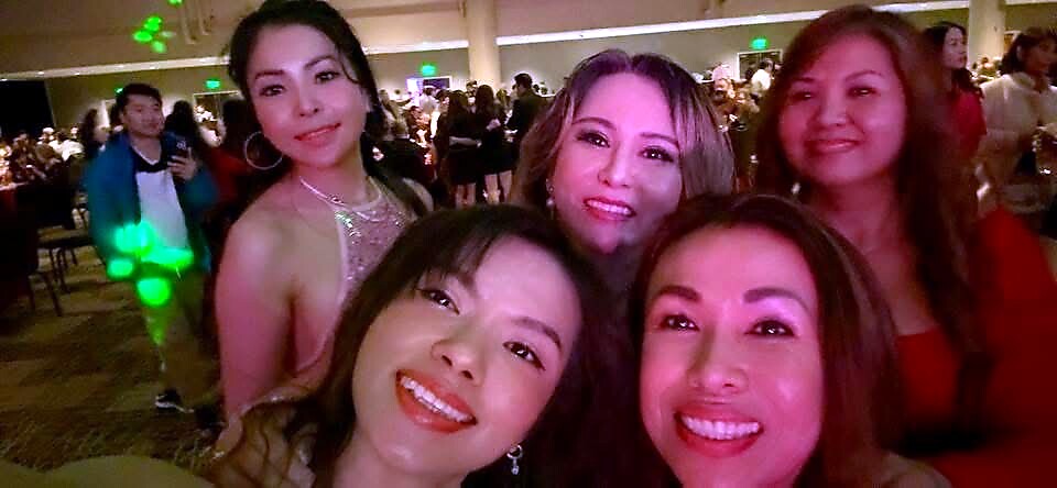 Five women smiling for a group selfie at a crowded party with colorful lighting in the background, facing the camera close up.”