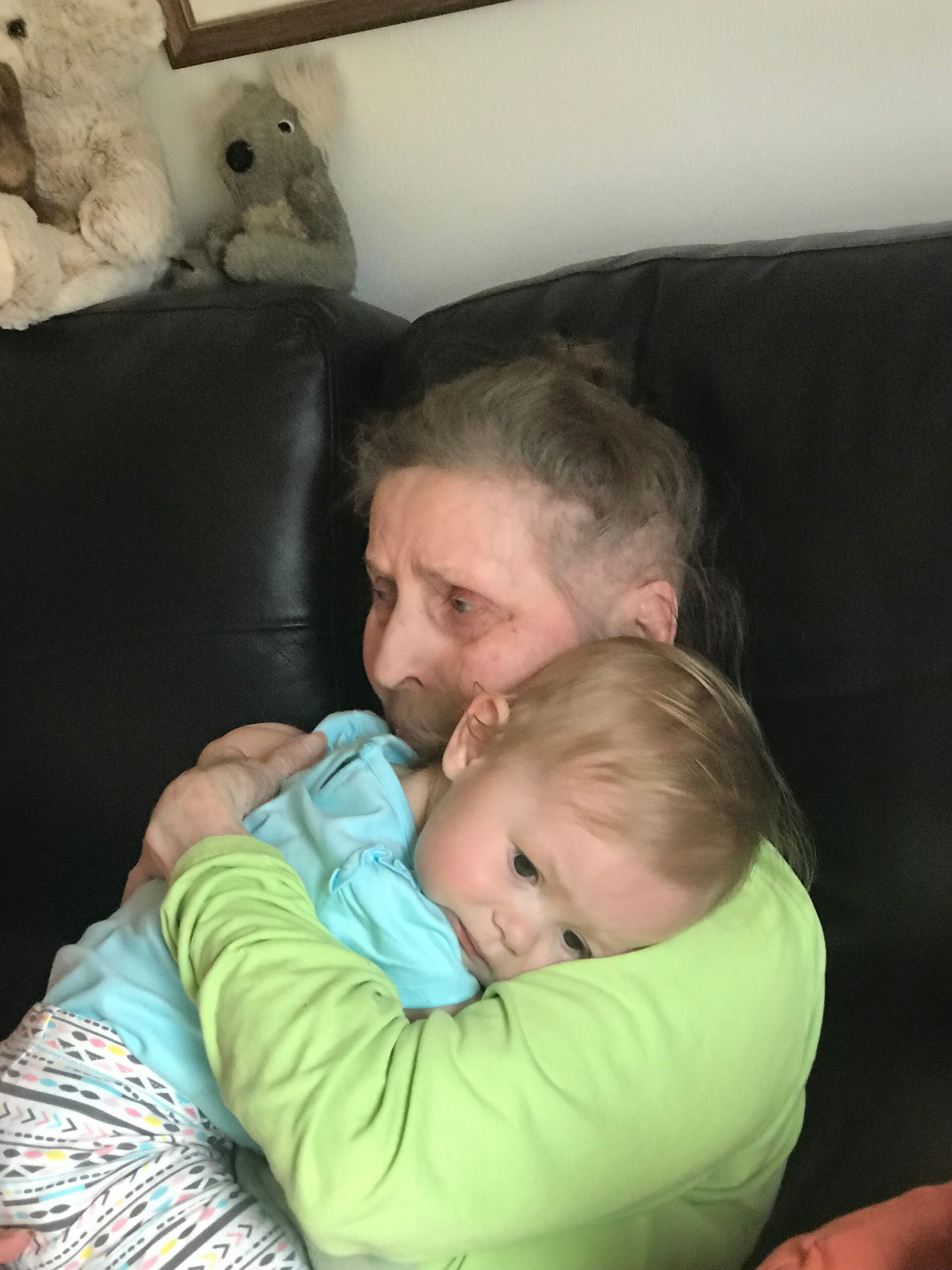 Older person cradling a baby on a dark leather couch, with a large stuffed bear in the background.