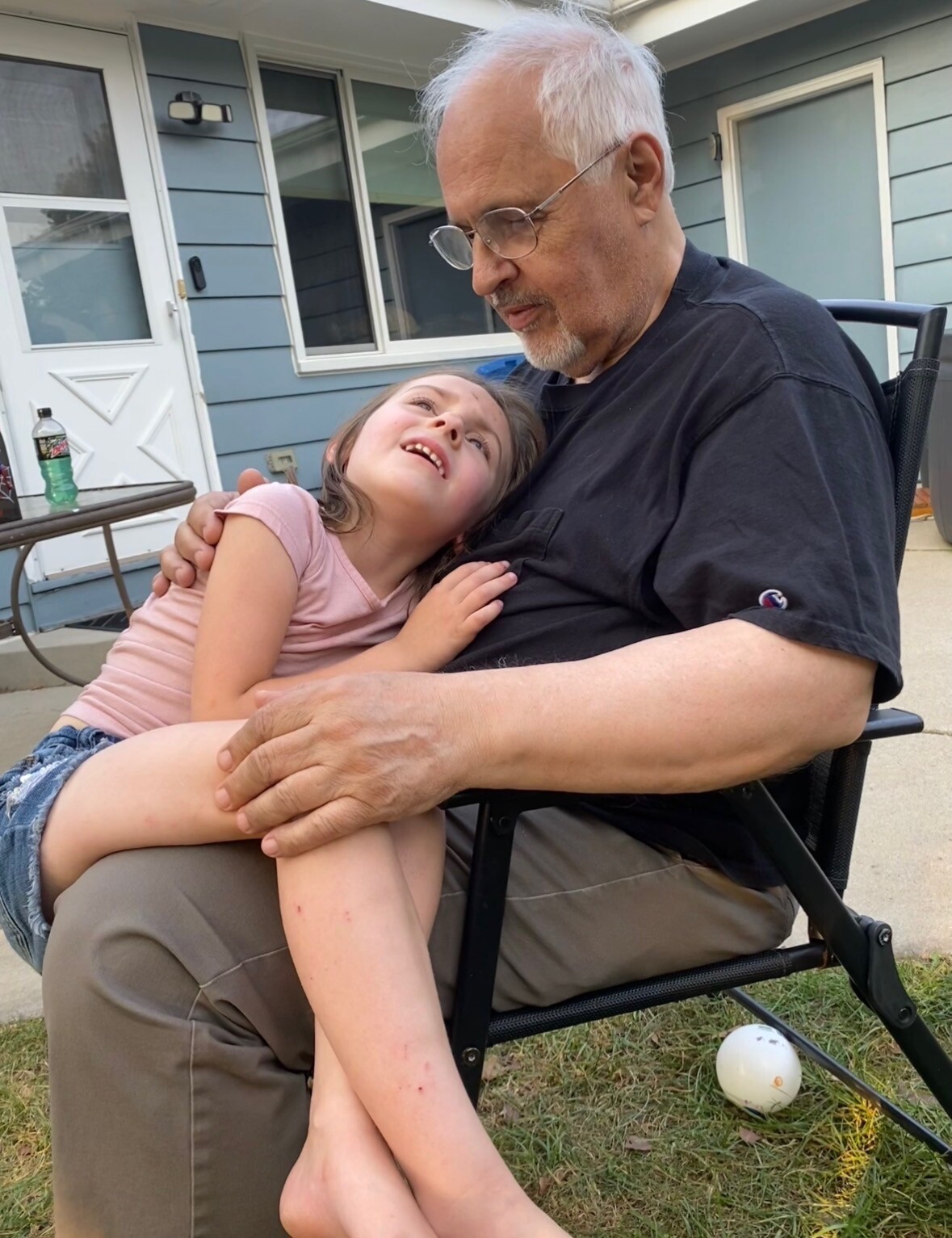 Older man with white hair and glasses sits in a reclining chair on a porch, smiling as a young girl rests in his lap and hugs his chest.