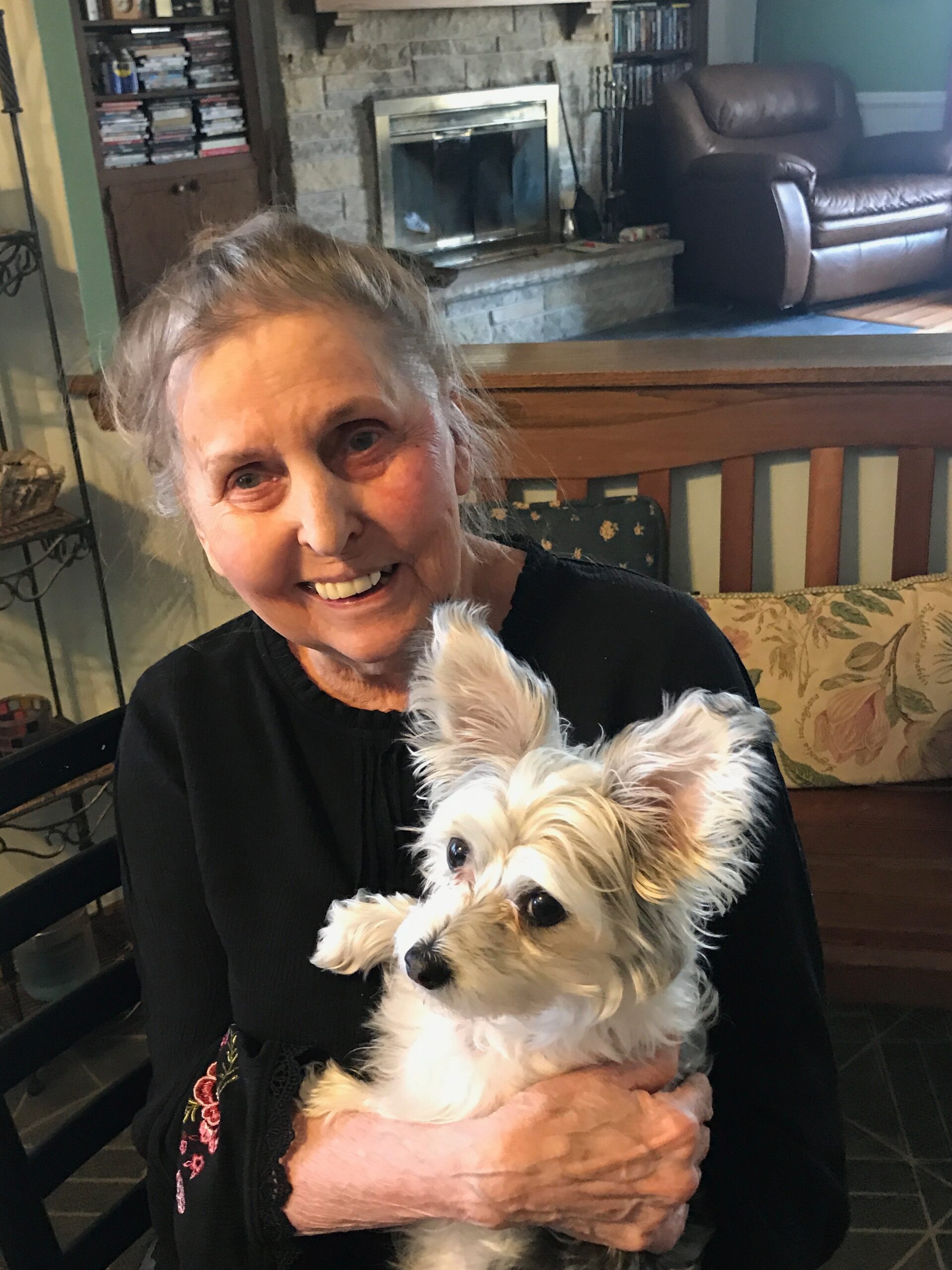 Smiling elderly woman in a black sweater holding a small fluffy white dog indoors in a living room with a stone fireplace behind them.