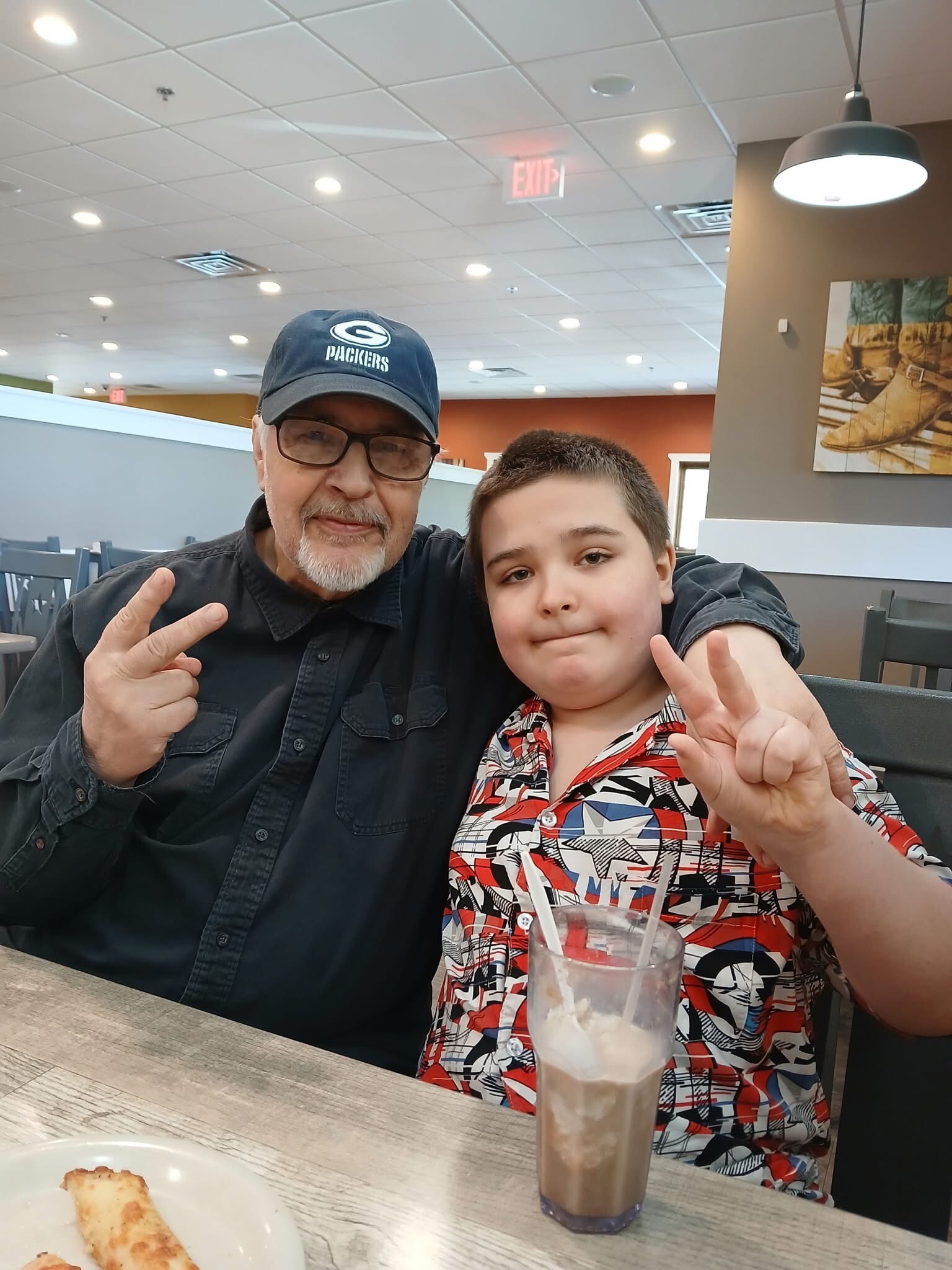 Two people posing for a selfie in a casual restaurant; man wearing a Packers cap and glasses, and a boy, both flashing peace signs. A tall chocolate milkshake is on the table in front of them.