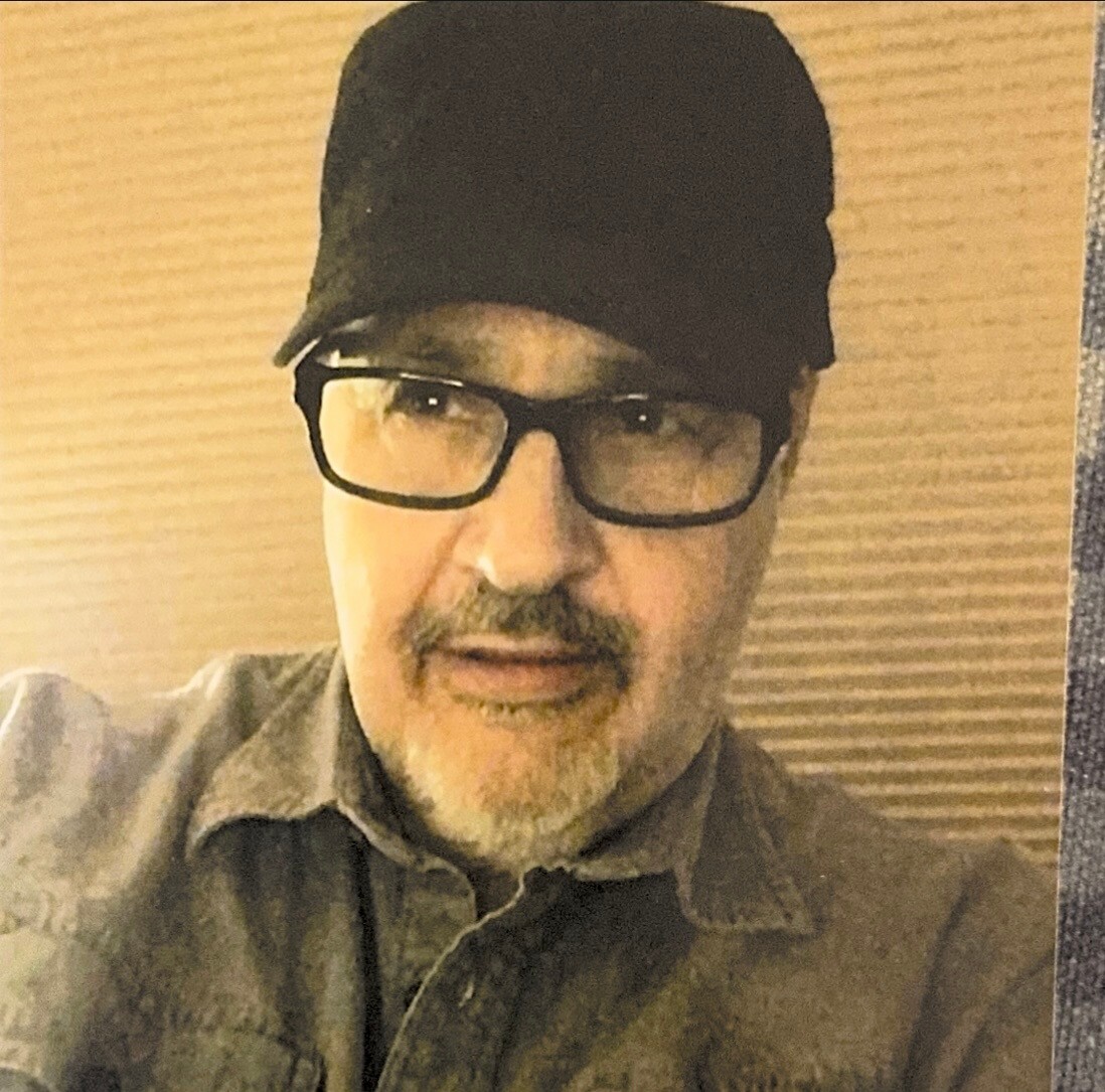 Portrait of a man with a gray beard wearing black-framed glasses and a black cap, facing the camera against a beige background.
