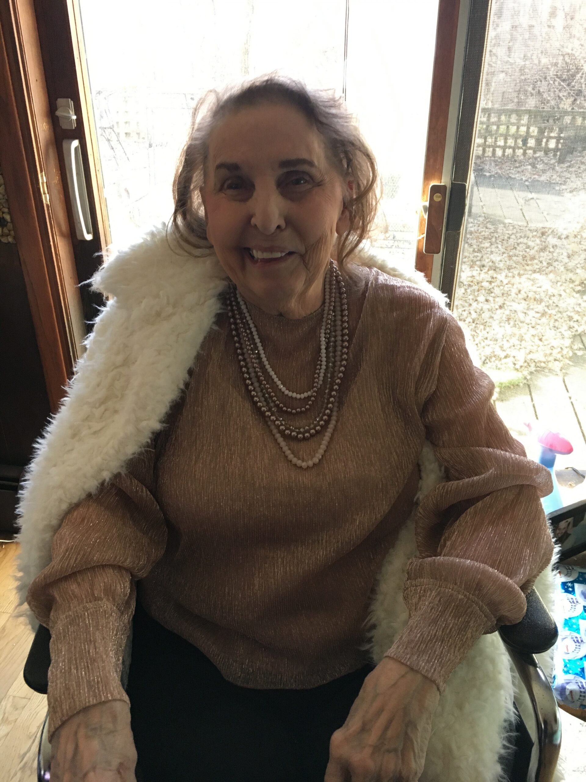 Smiling elderly woman in a brown shimmery top with layered pearl necklaces and a white fluffy scarf, seated by a window.