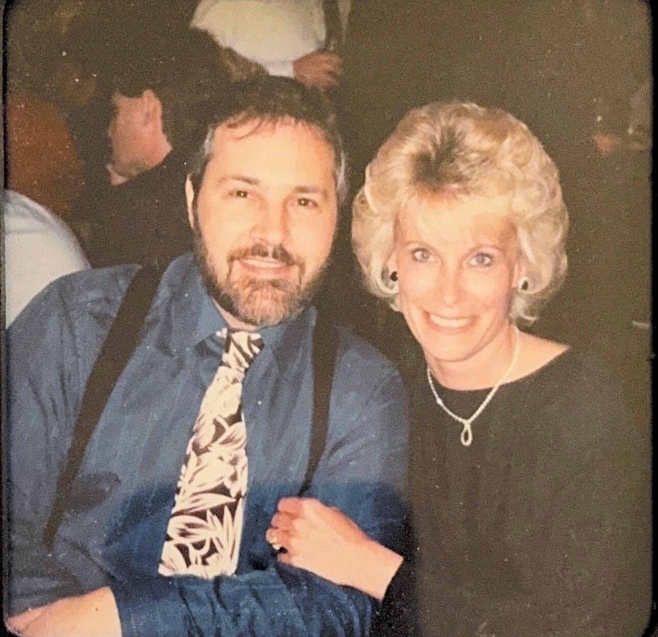 Smiling man with a beard in a blue shirt and tropical tie posing with a woman in a black dress at a social event.