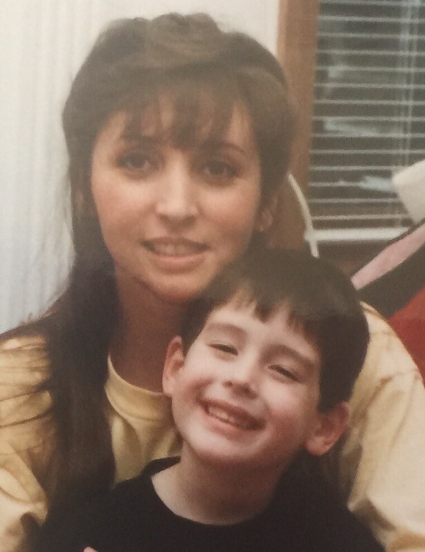 Woman with long brown hair and bangs gently embracing a smiling boy indoors.