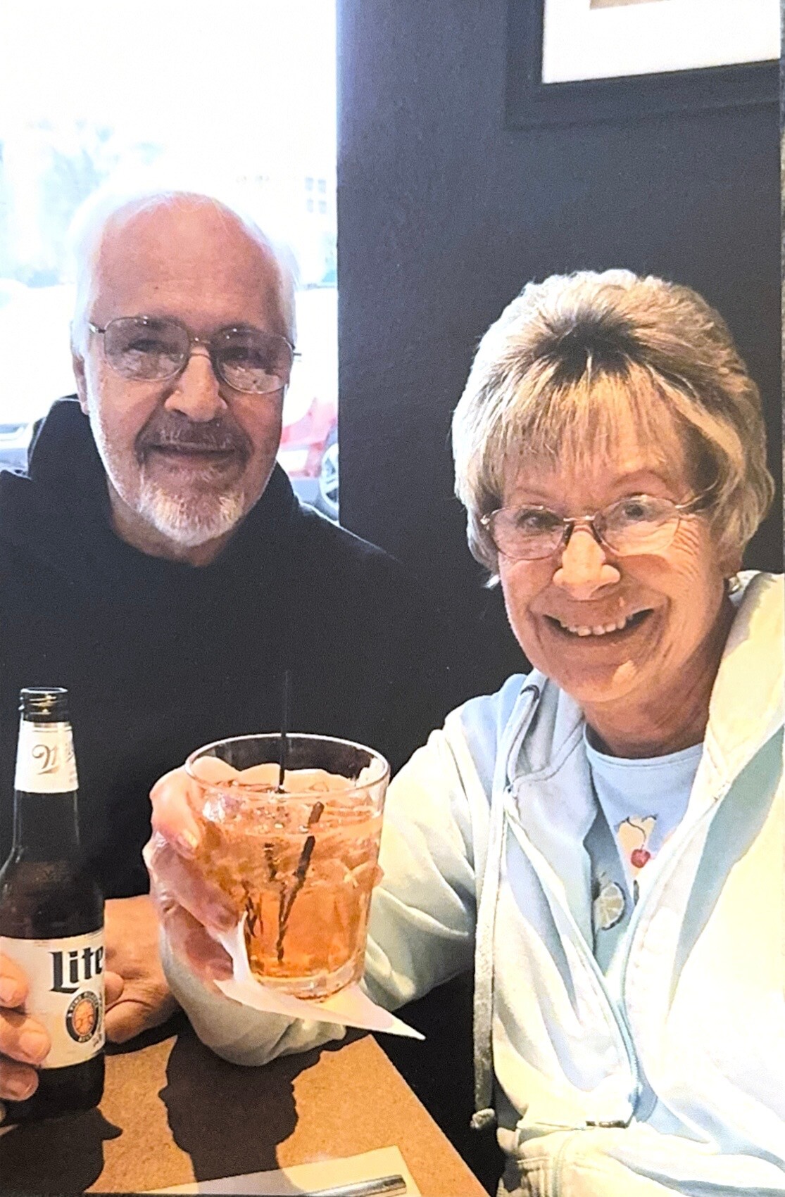 Two smiling seniors sit at a restaurant table, sharing drinks—a beer bottle on the left and a pink cocktail in the center