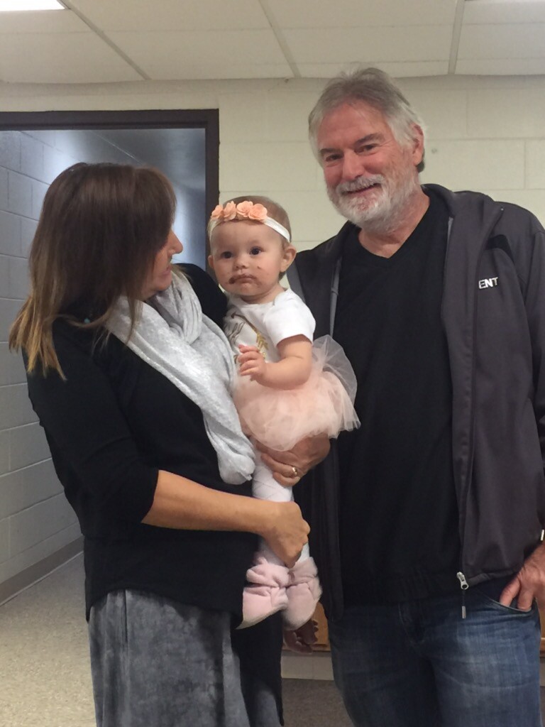 Woman holding a baby girl in a pink tutu while a smiling man stands beside them in a hallway.