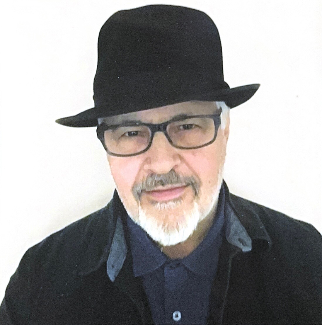 Portrait of an older man with gray beard wearing a black fedora, glasses, and a dark jacket, facing the camera.