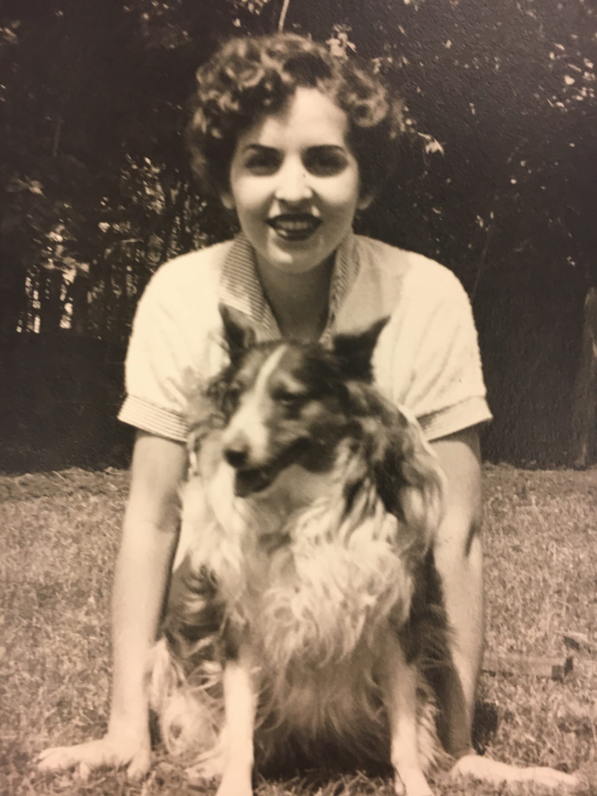 Smiling woman outdoors kneeling or sitting with a long-haired fluffy dog in front of her on the grass (black-and-white photo).