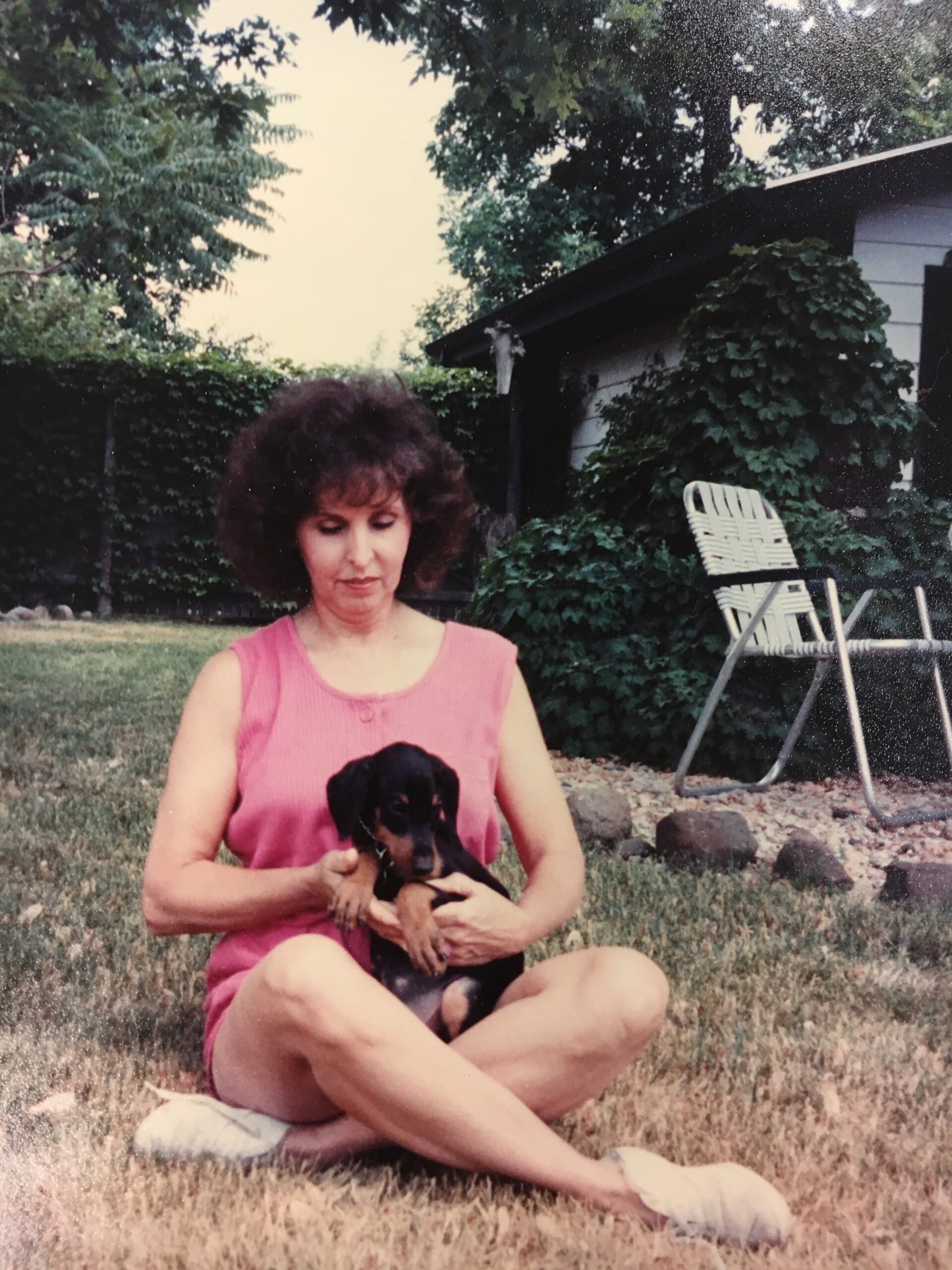 Woman in a pink sleeveless dress sits on the grass, cradling a small black puppy outdoors.