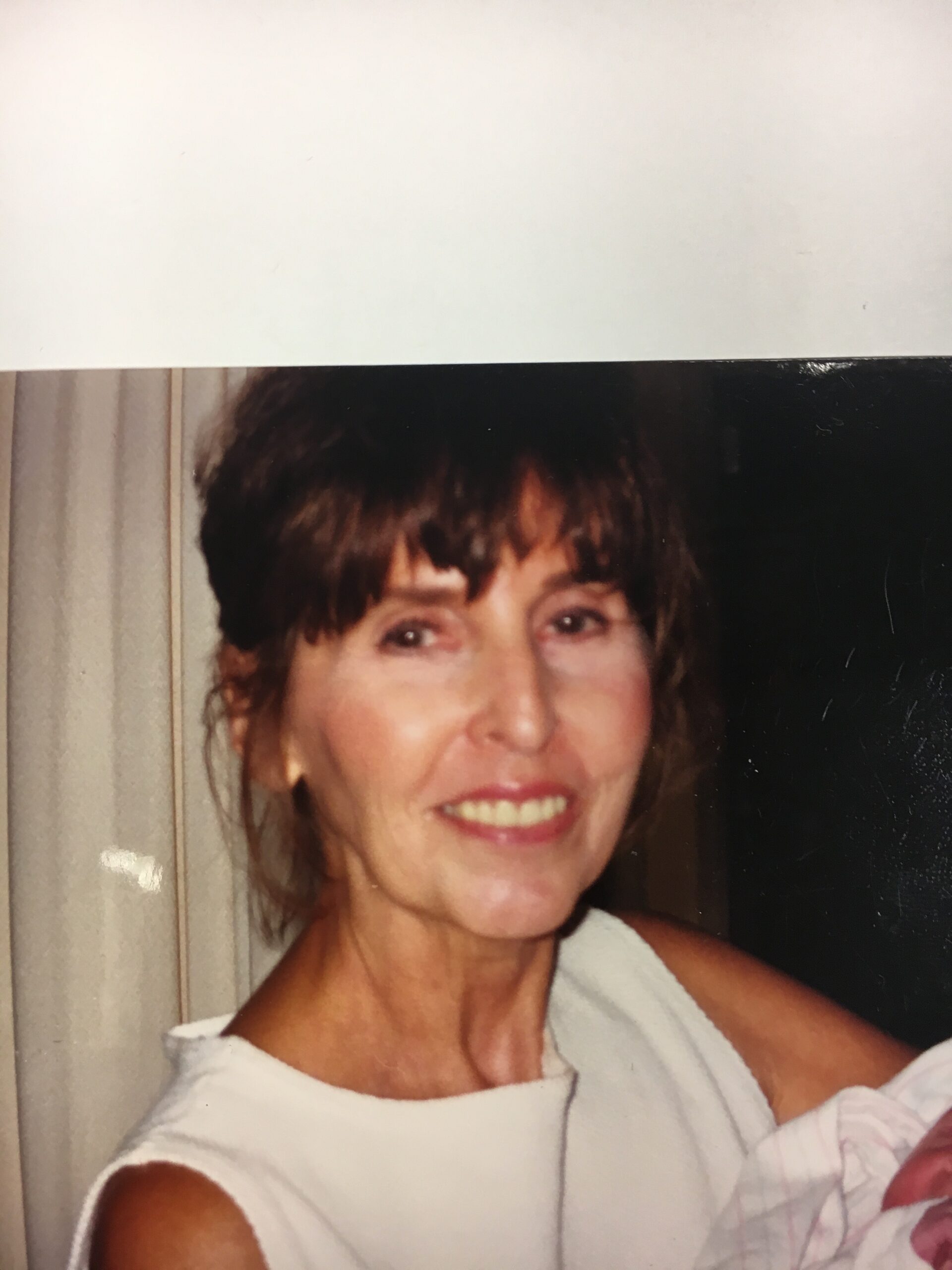 Smiling woman with short brown hair wearing a white sleeveless top, posing indoors.