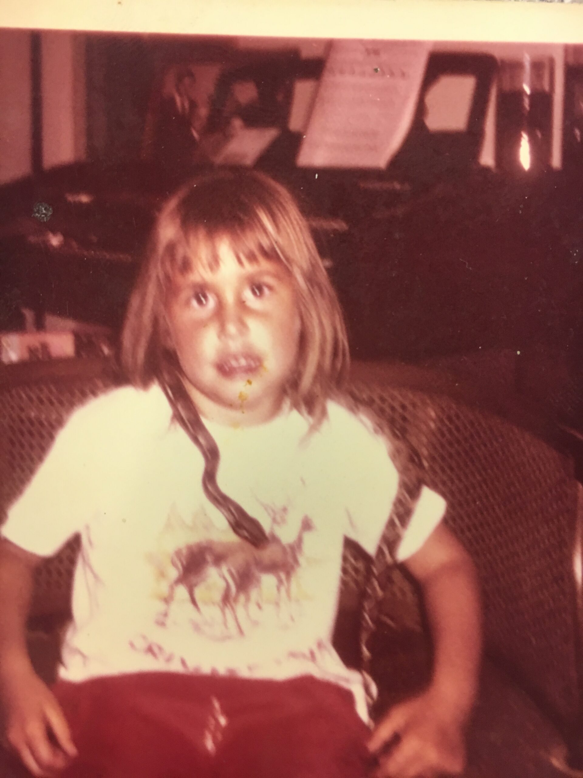 A young child with light brown hair sits on a red couch indoors, wearing a white T-shirt with a graphic design.