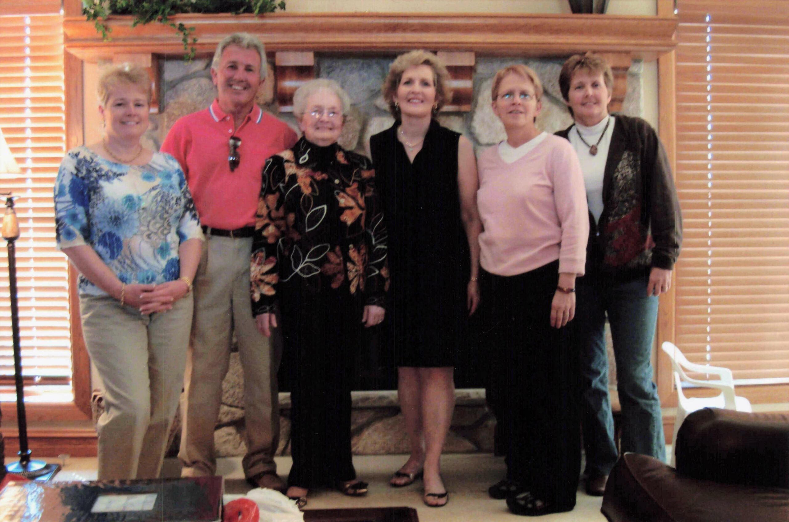 Group of six adults posing together indoors in front of a stone fireplace, smiling at the camera.