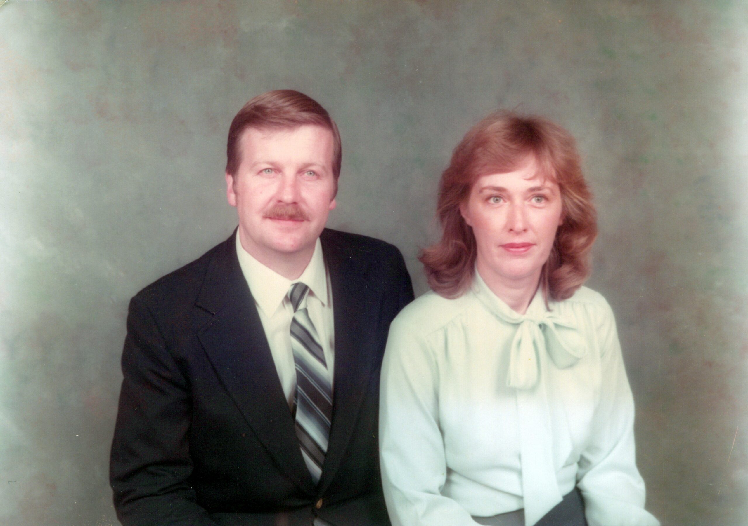 Man in a dark suit with striped tie sits beside a red-haired woman in a light blue blouse for a studio portrait.