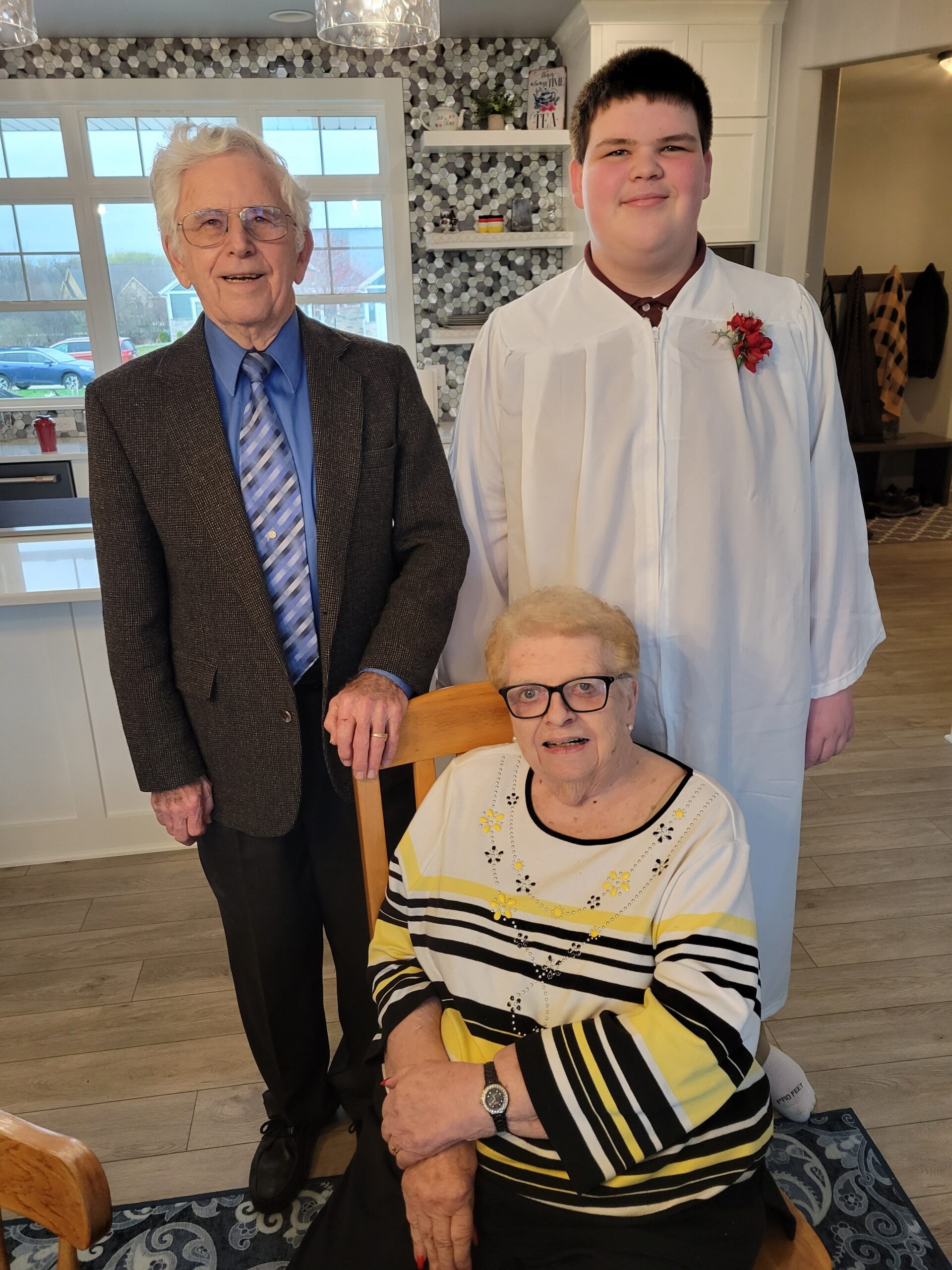 Elderly woman seated at a kitchen table in a striped sweater, with two men standing beside her.