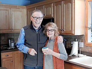 Smiling older couple with arms around each other standing in a cozy kitchen; the woman holds a cup.