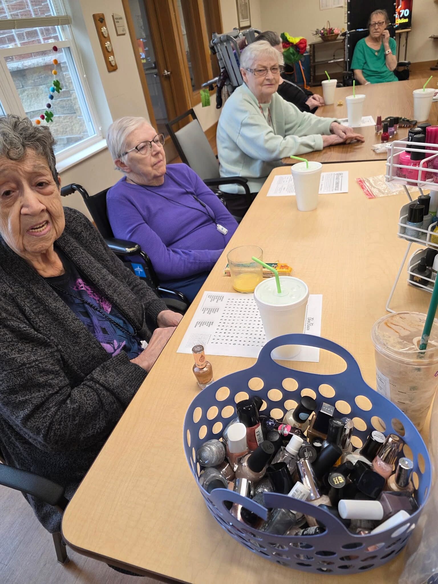 Group of elderly women seated around a table in a activity room, manicure supplies in a blue basket and coffee cups on the table, suggesting a social nail-painting or spa activity.