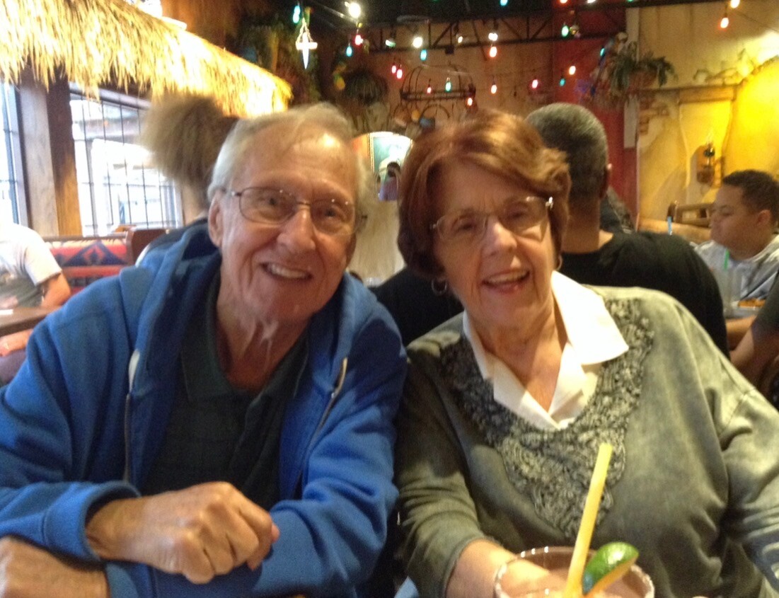 Smiling elderly man in a blue jacket and an elderly woman with glasses sit at a colorful restaurant table under string lights, posing for the camera with drinks nearby.