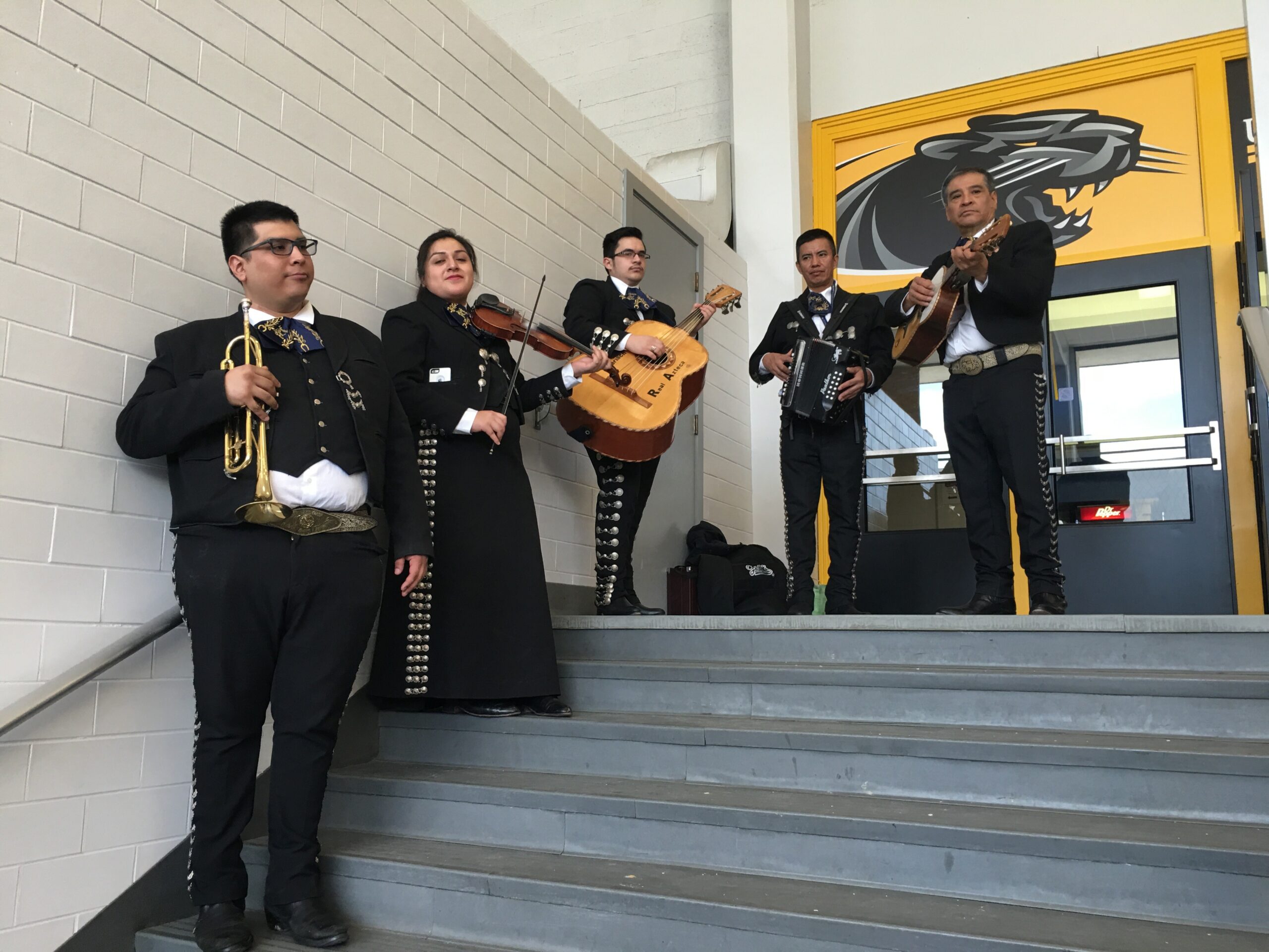A group of mariachi musicians in traditional black outfits with silver trim perform on a short staircase indoors, holding trumpets, violin, guitar, and accordion.