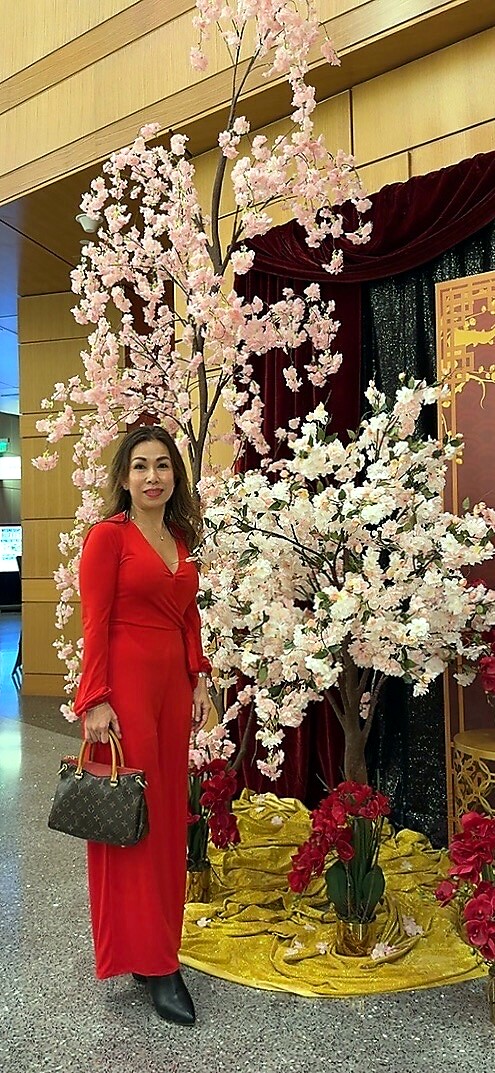 Woman in a red outfit posing beside a pink cherry blossom display indoors, holding a dark handbag.