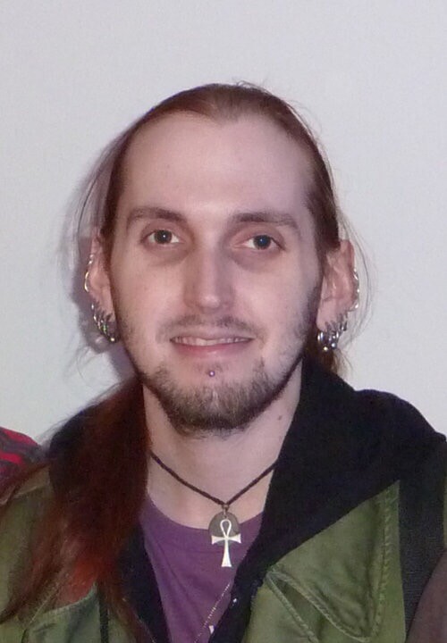 Portrait of a man with long reddish-brown hair, multiple ear piercings, and a goatee, wearing a green jacket and an ankh necklace.