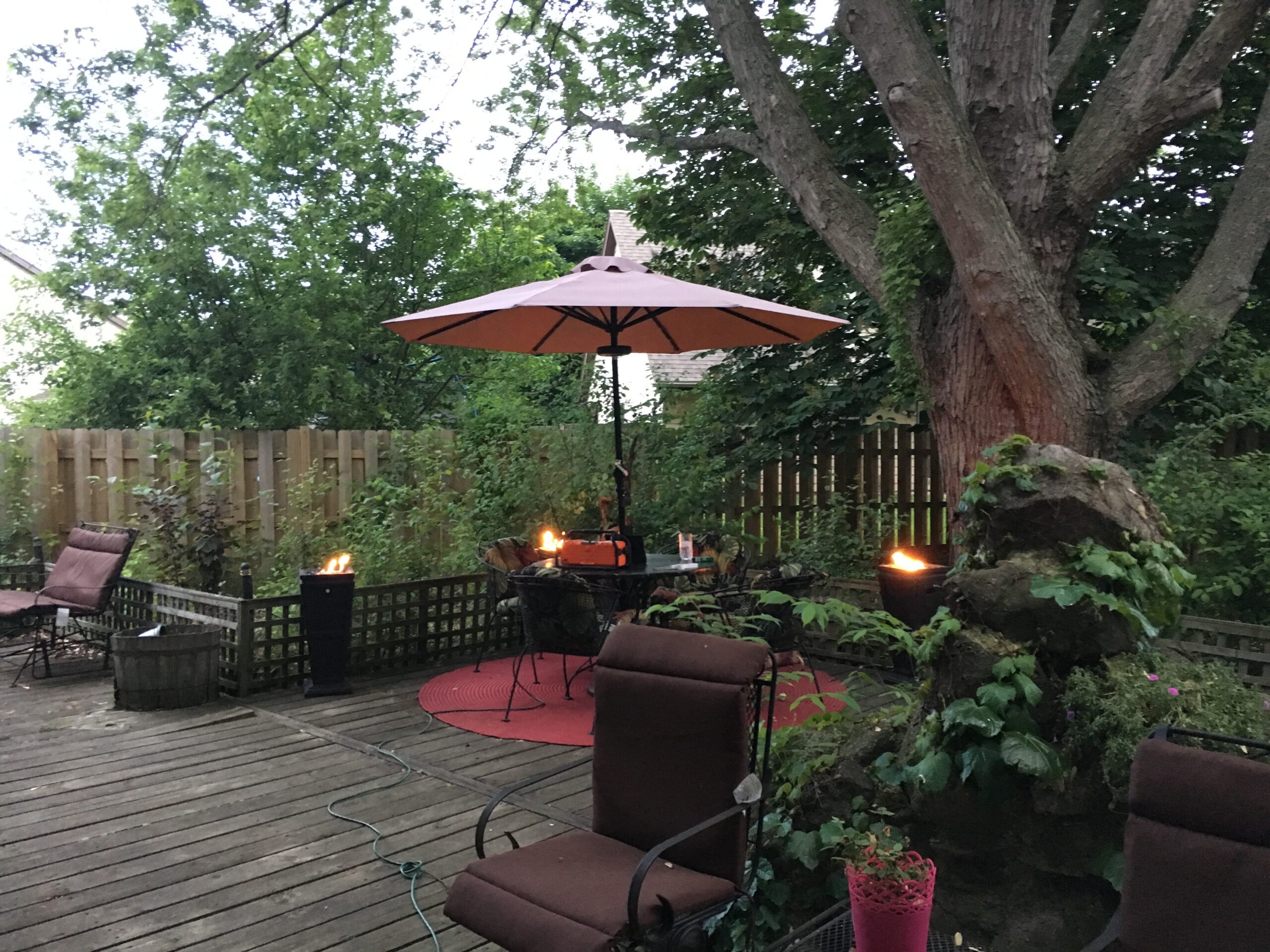 Outdoor backyard patio with a pink umbrella, cushioned chairs, and a tall tree in the background by a wooden fence.