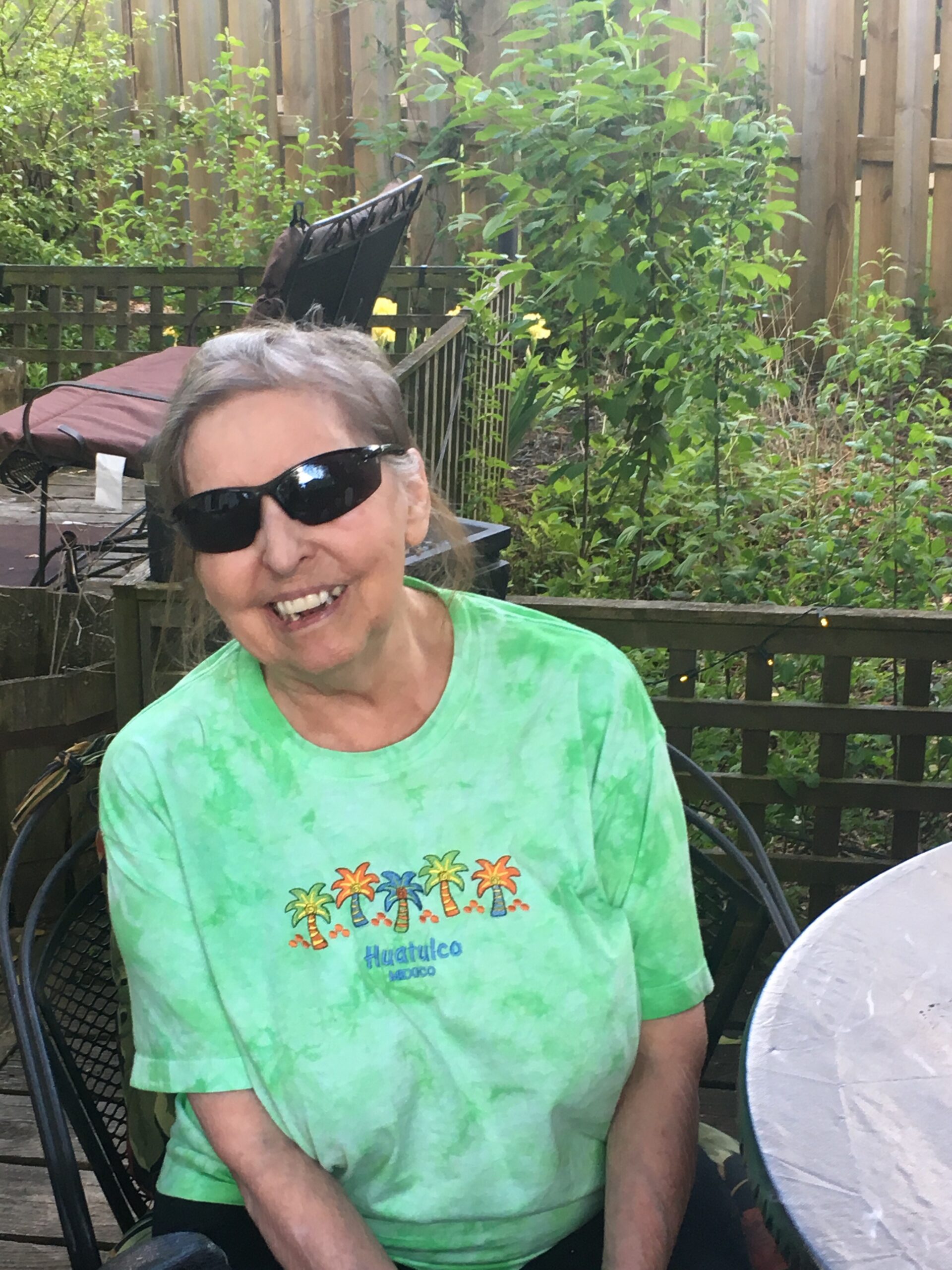 Smiling woman wearing black sunglasses and a green tie-dye shirt, seated in a backyard with a wooden fence and plants behind her