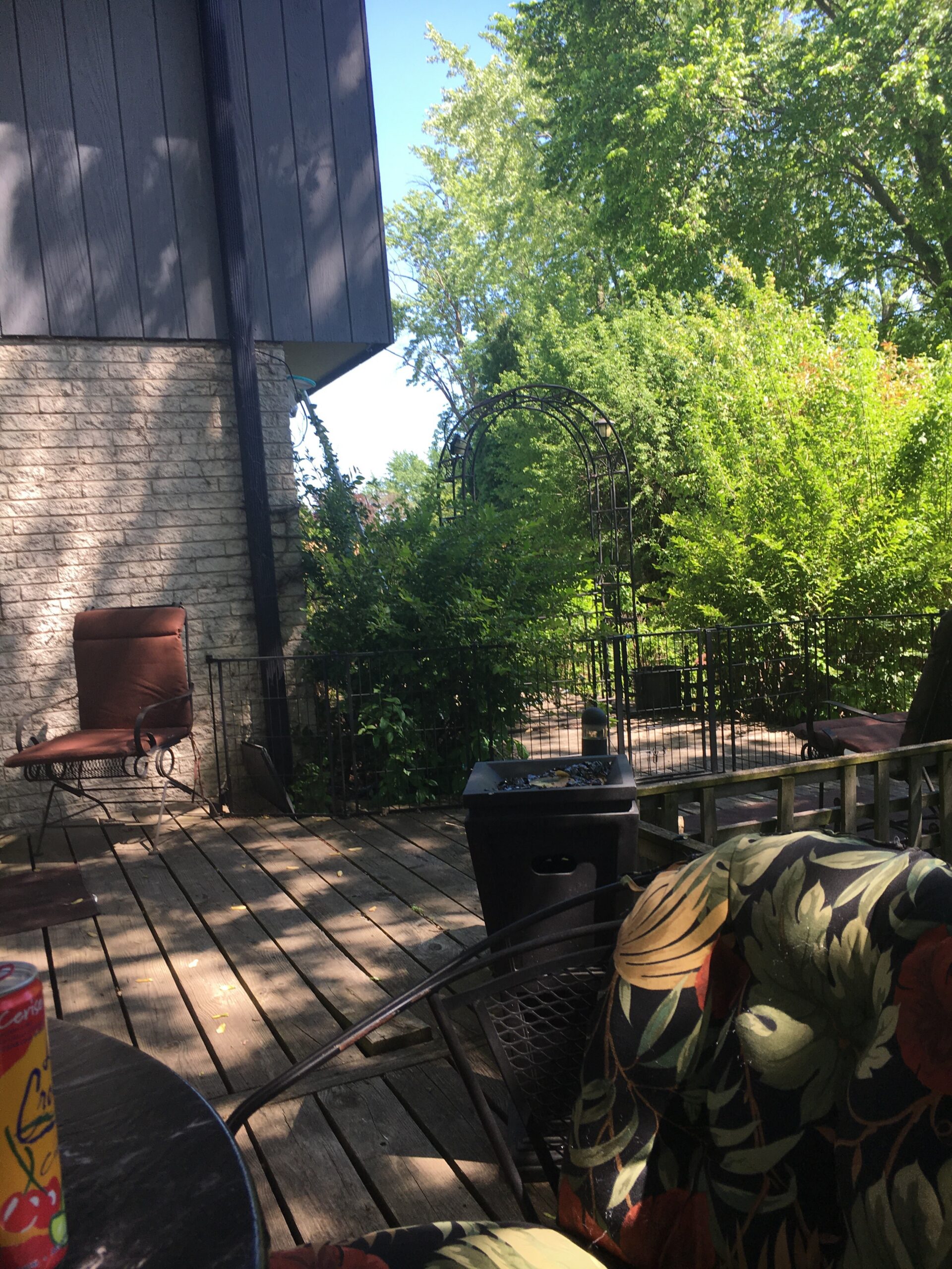Wooden deck patio beside a brick wall, with cushioned chairs, a metal table, and a grill, surrounded by lush green trees and a black metal fence.