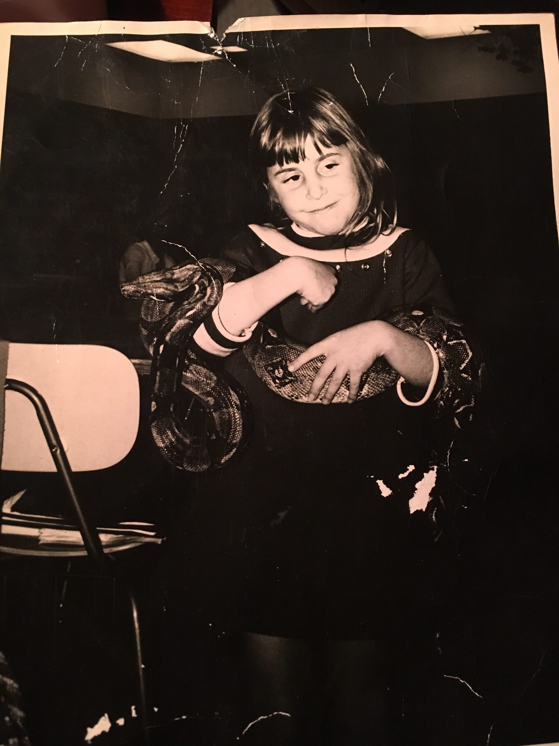 Young girl in a dark dress sits with arms crossed, a calm, shy smile on her face. The photo is damaged with creases around the edges.