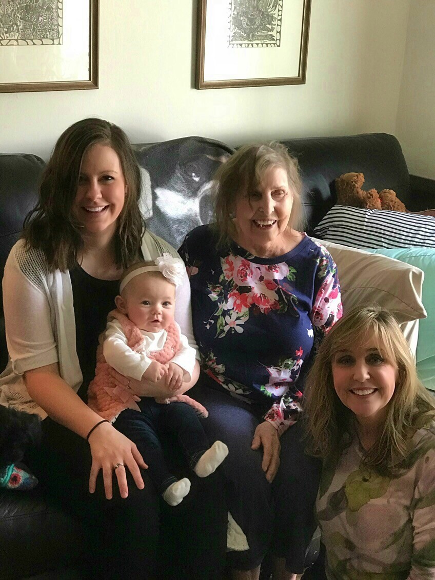 Three generations of women with a baby posing on a dark sofa, smiling in a living room setting.