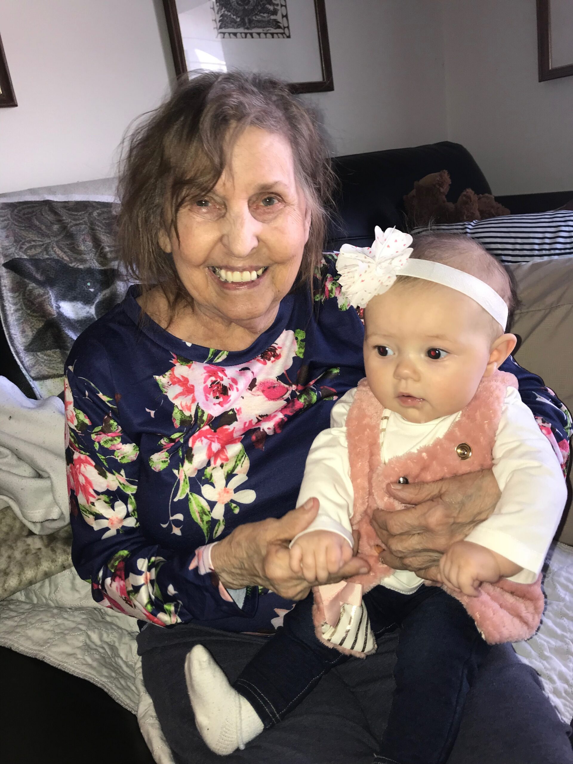 Smiling elderly woman with floral blouse sits on a couch, hugging a baby doll in a pink fuzzy vest and white headband.