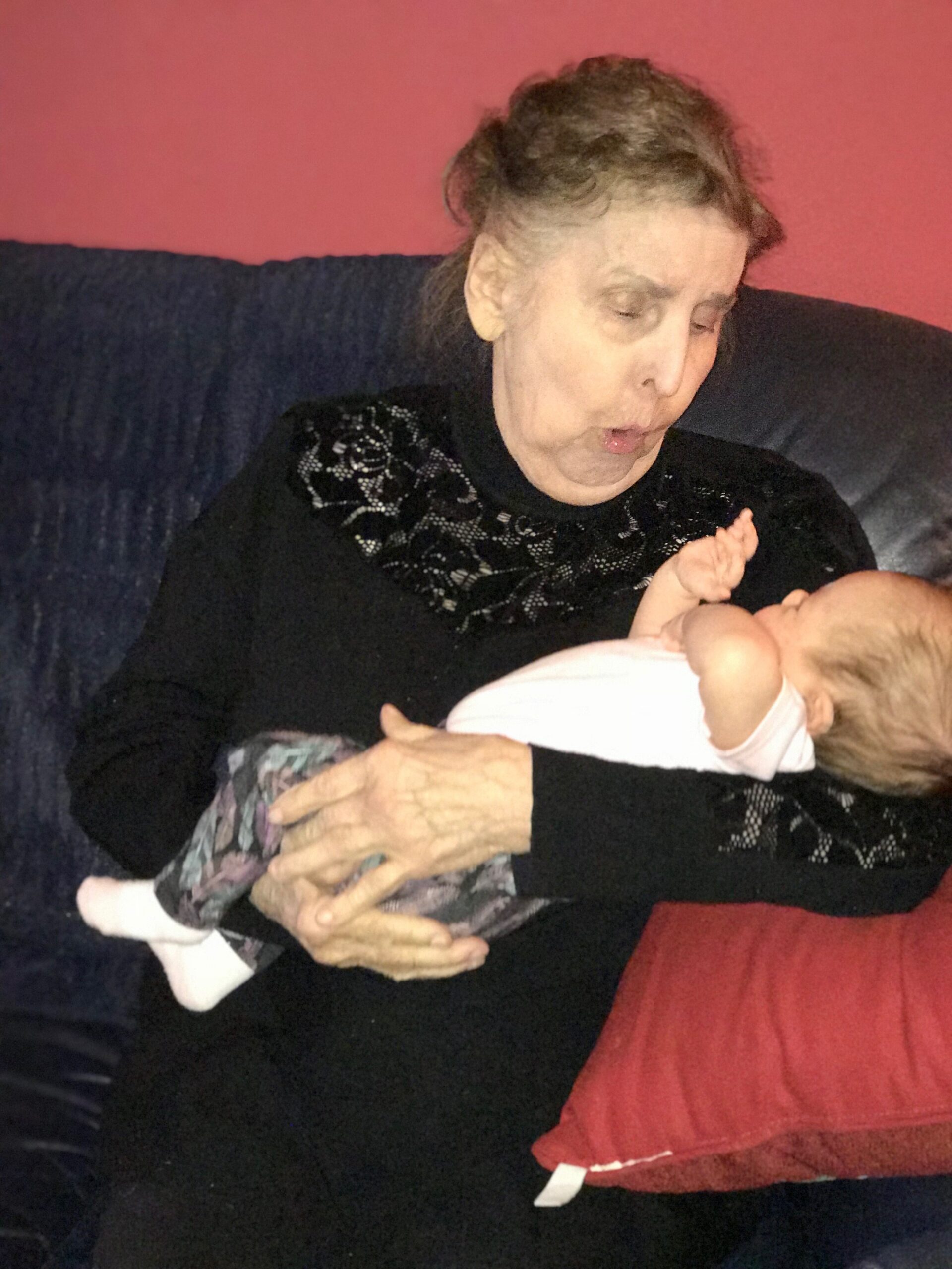 Elderly woman in a black lace top cradles a newborn baby on a dark sofa with a pink wall behind them.