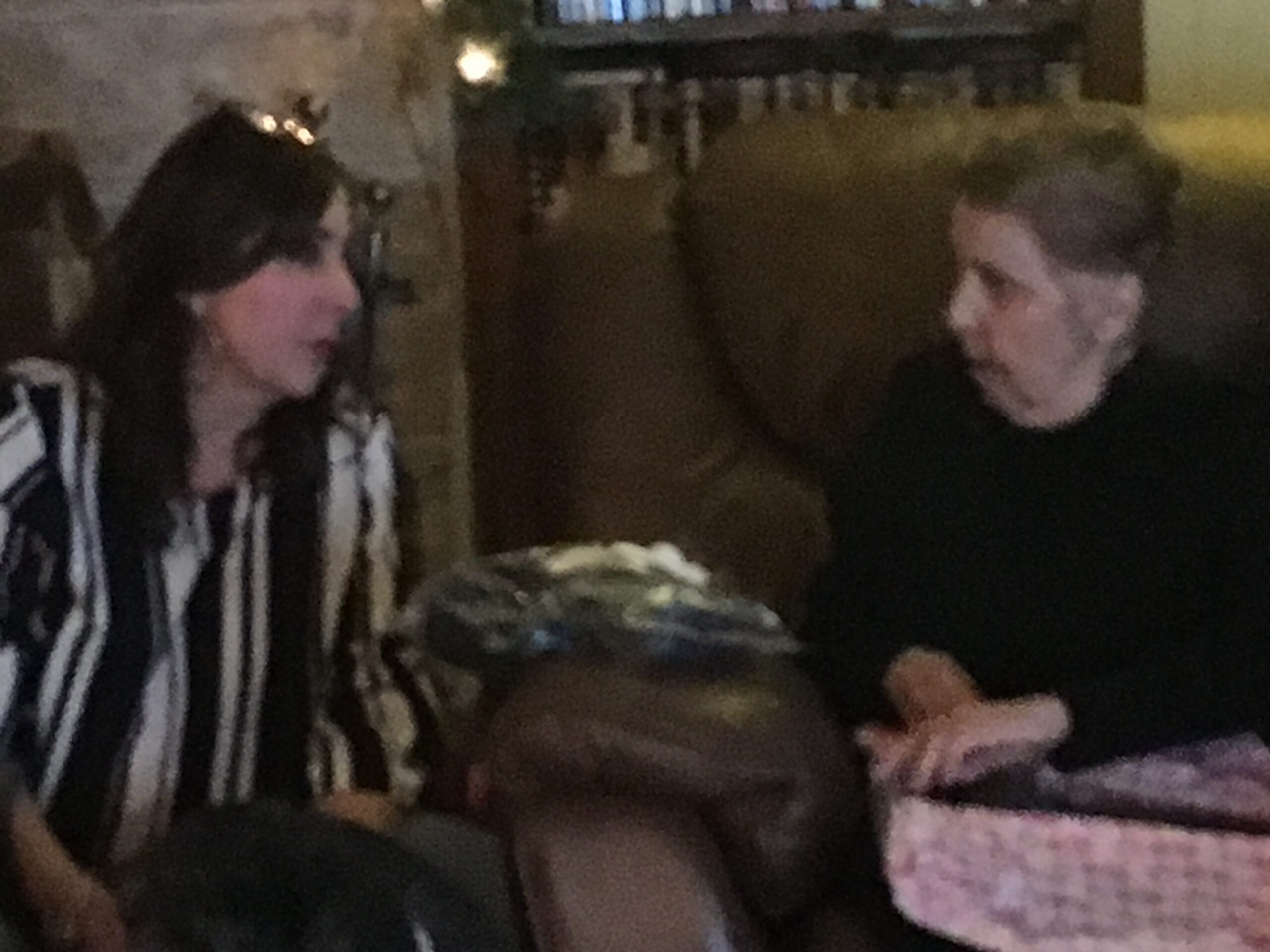 Two people seated at a dining table in a dim room: a woman with dark hair wearing a black-and-white striped jacket on the left and a man in dark clothing on the right.