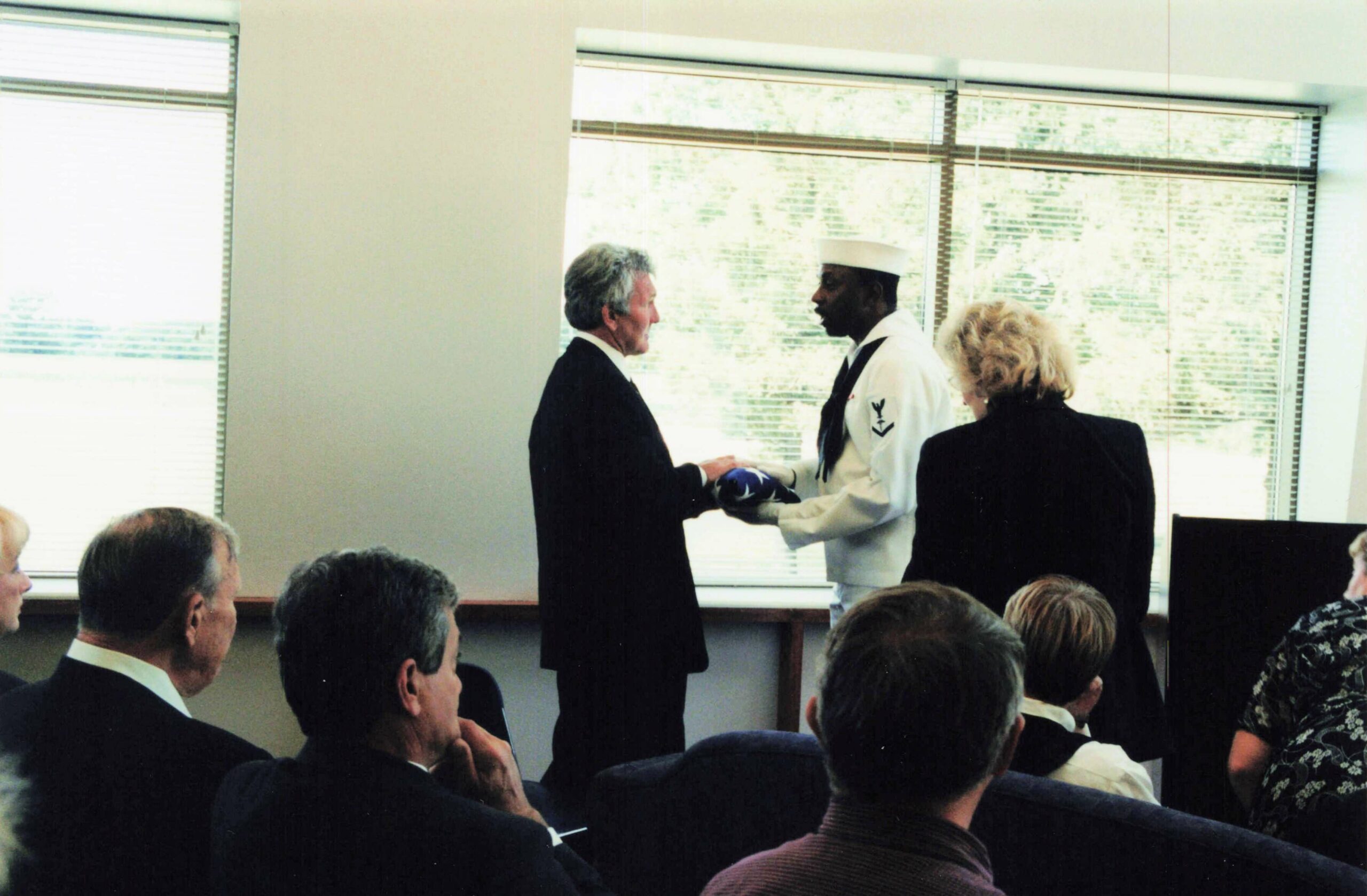 A uniformed service member presents a folded flag to an older man in a dark suit in a conference room ceremony, while others look on.