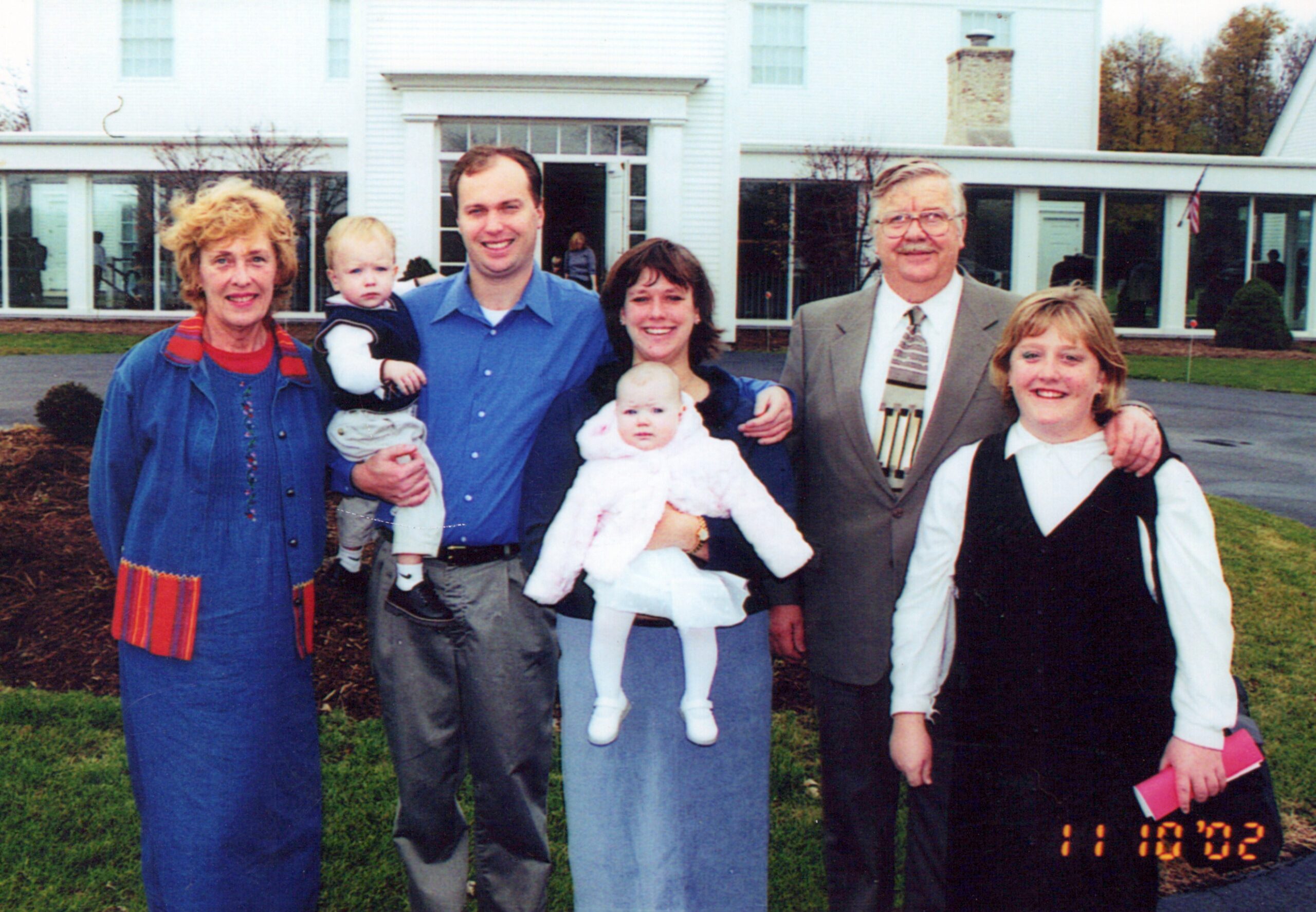 Group of seven people posing outdoors in front of a white building, two babies in arms, all smiling at the camera.