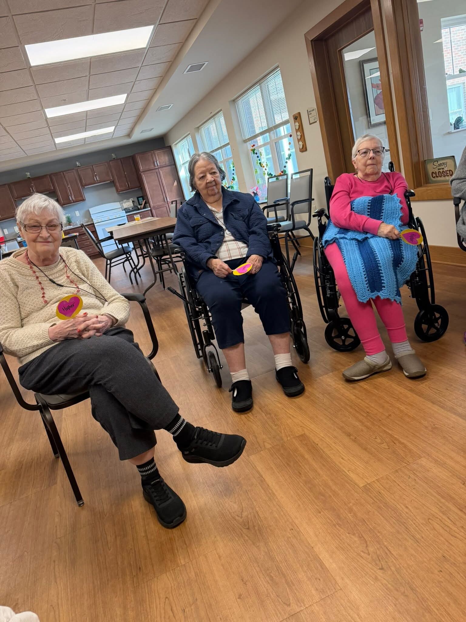 Three elderly women in wheelchairs sit in a community room, holding heart-shaped notes, smiling at the camera.