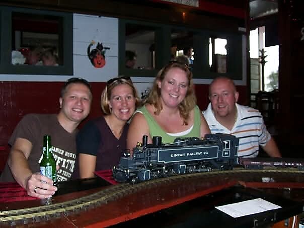 Four adults at a bar smiling for the camera, with a blue model train on a curved track on the counter (one man holds a beer).