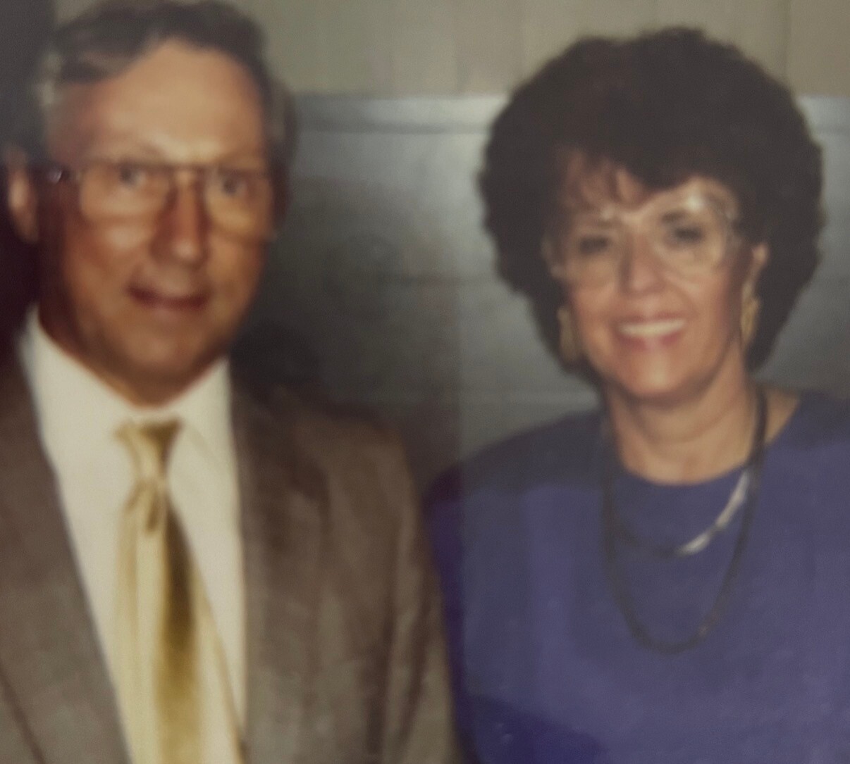Smiling older man in a tan suit with a yellow tie standing next to a smiling older woman in a royal blue top.
