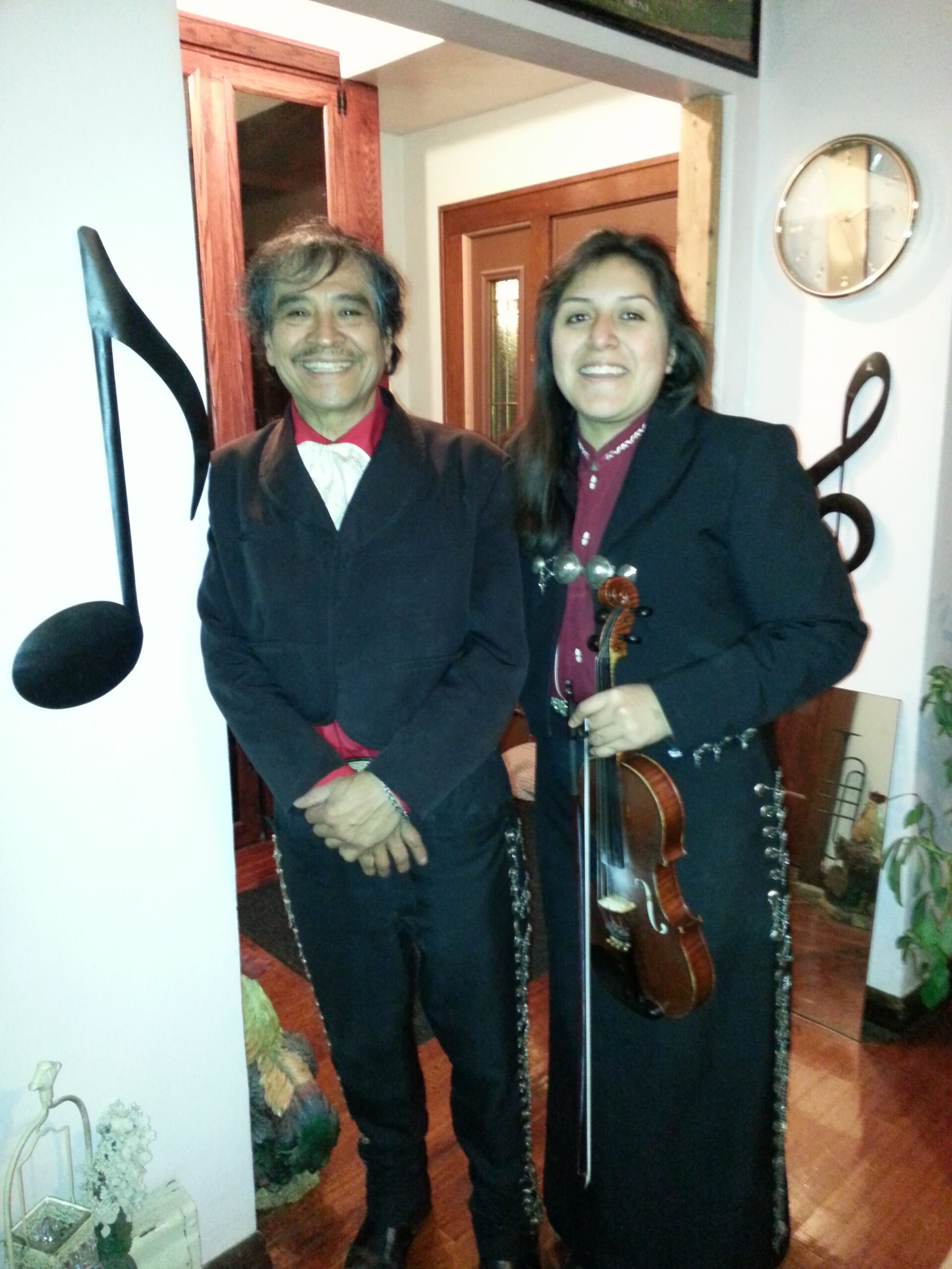 Two musicians stand indoors in a doorway; man in a red shirt and bow tie smiles, woman in a blazer holds a violin and smiles.