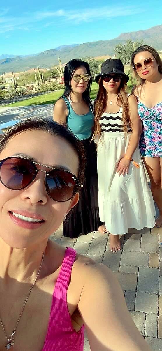 Four women posing for a sunny outdoor selfie, with mountains in the background; the woman in the foreground wears a pink top and sunglasses.