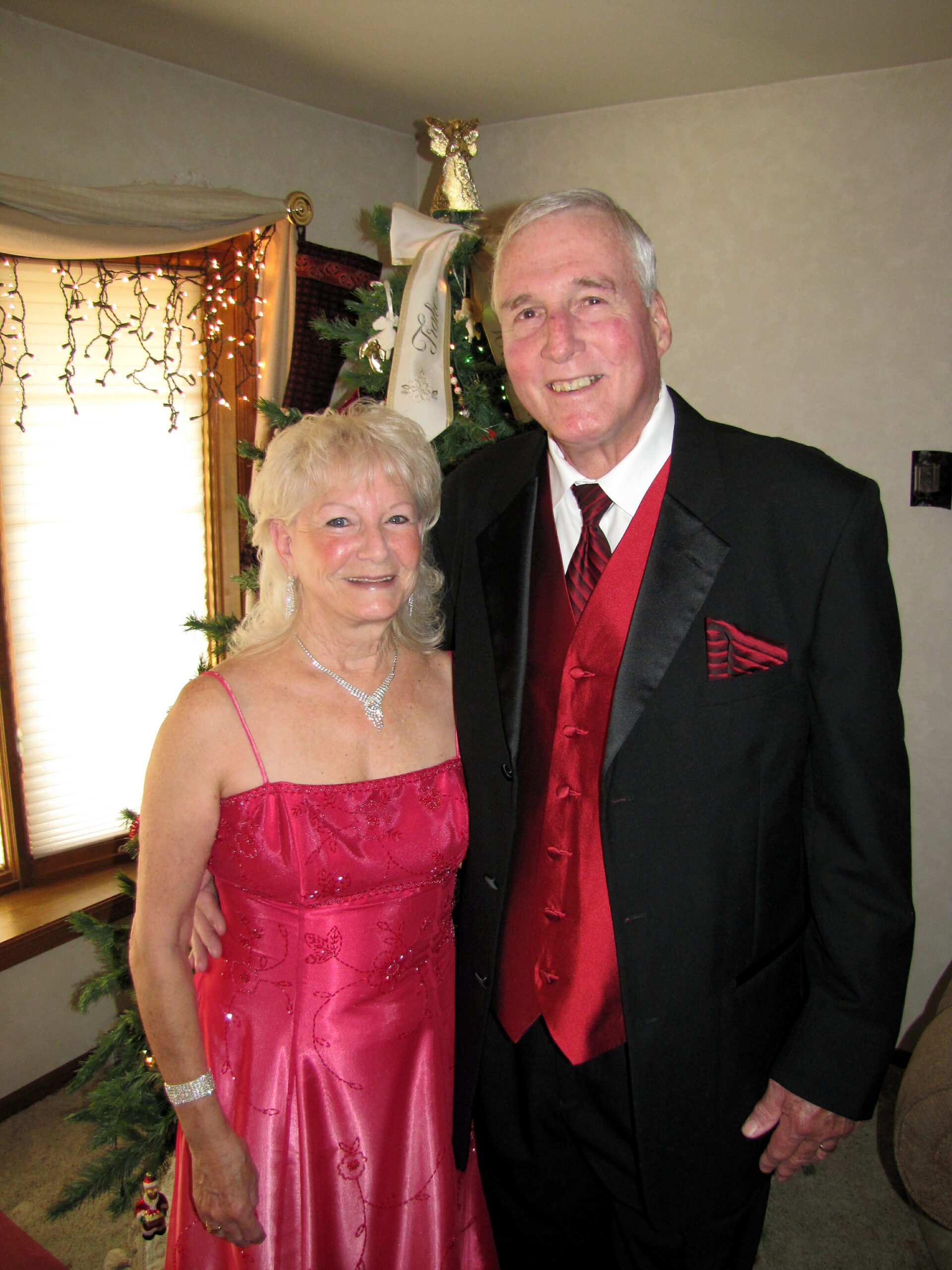 Smiling elderly couple in formal attire posing by a decorated Christmas tree indoors.