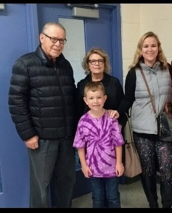 Four people pose for a photo in a school hallway: an older man on the left, an older woman behind the boy, a smiling woman on the right, and a young boy in a purple tie-dye shirt in front.