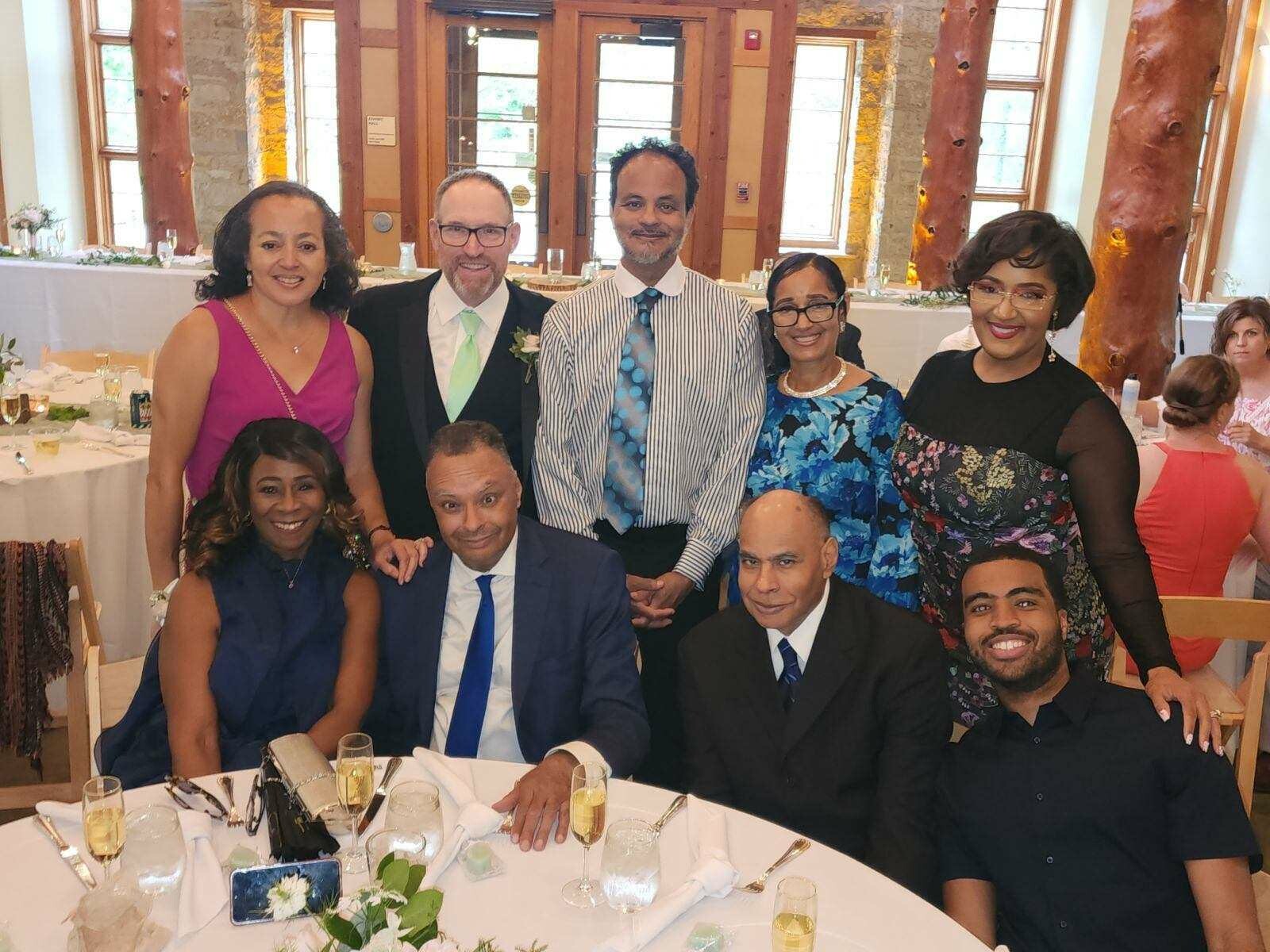Group of eight adults at a wedding reception posing for a photo around a table with champagne flutes on it.