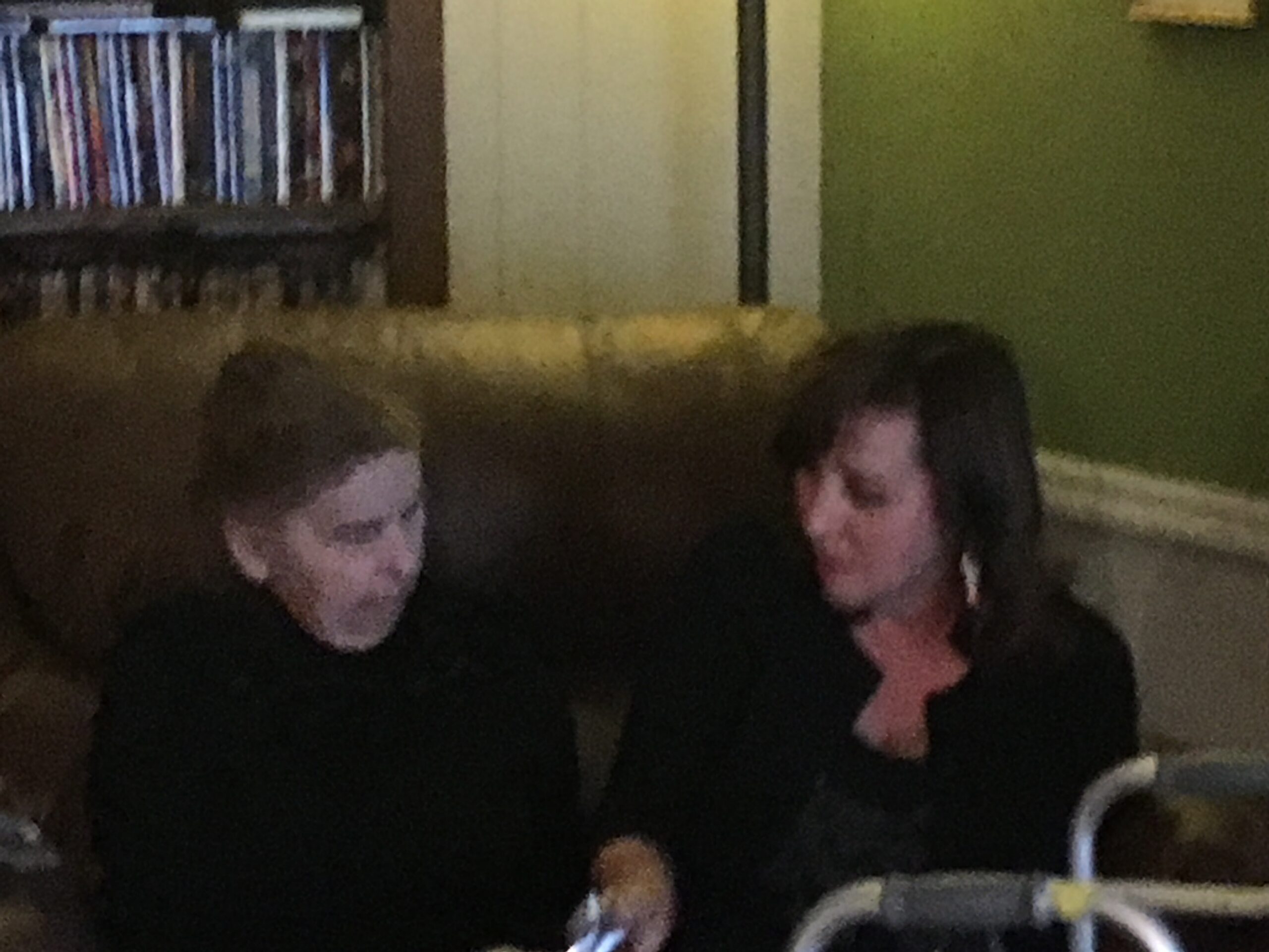 Two people in black shirts pose for a photo indoors, with a bookshelf along a beige wall on the left and a green wall on the right.